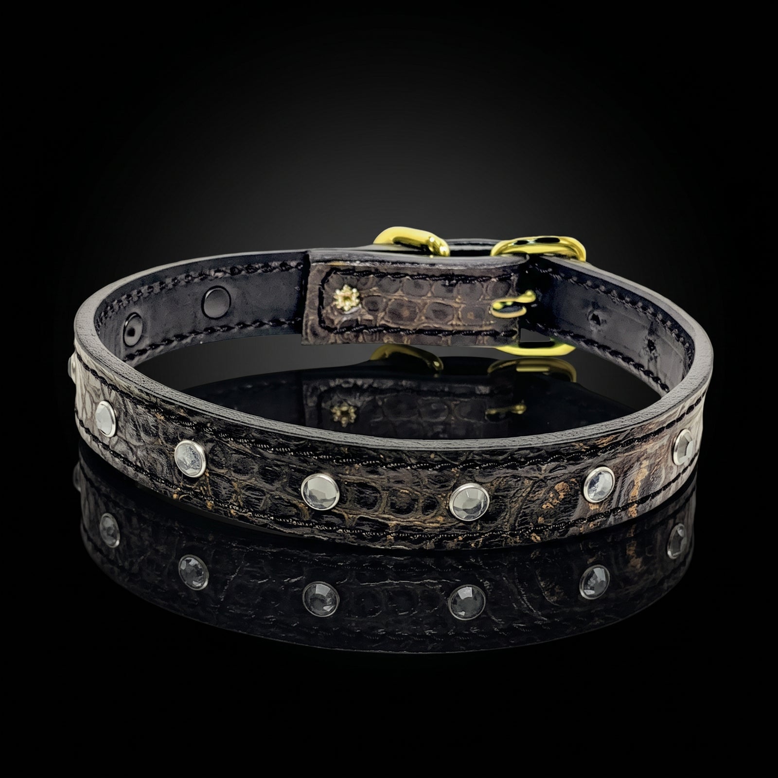 A dark brown crocodile-embossed leather collar with gold-tone hardware and silver-tone studs, displayed on a reflective black surface against a black background.