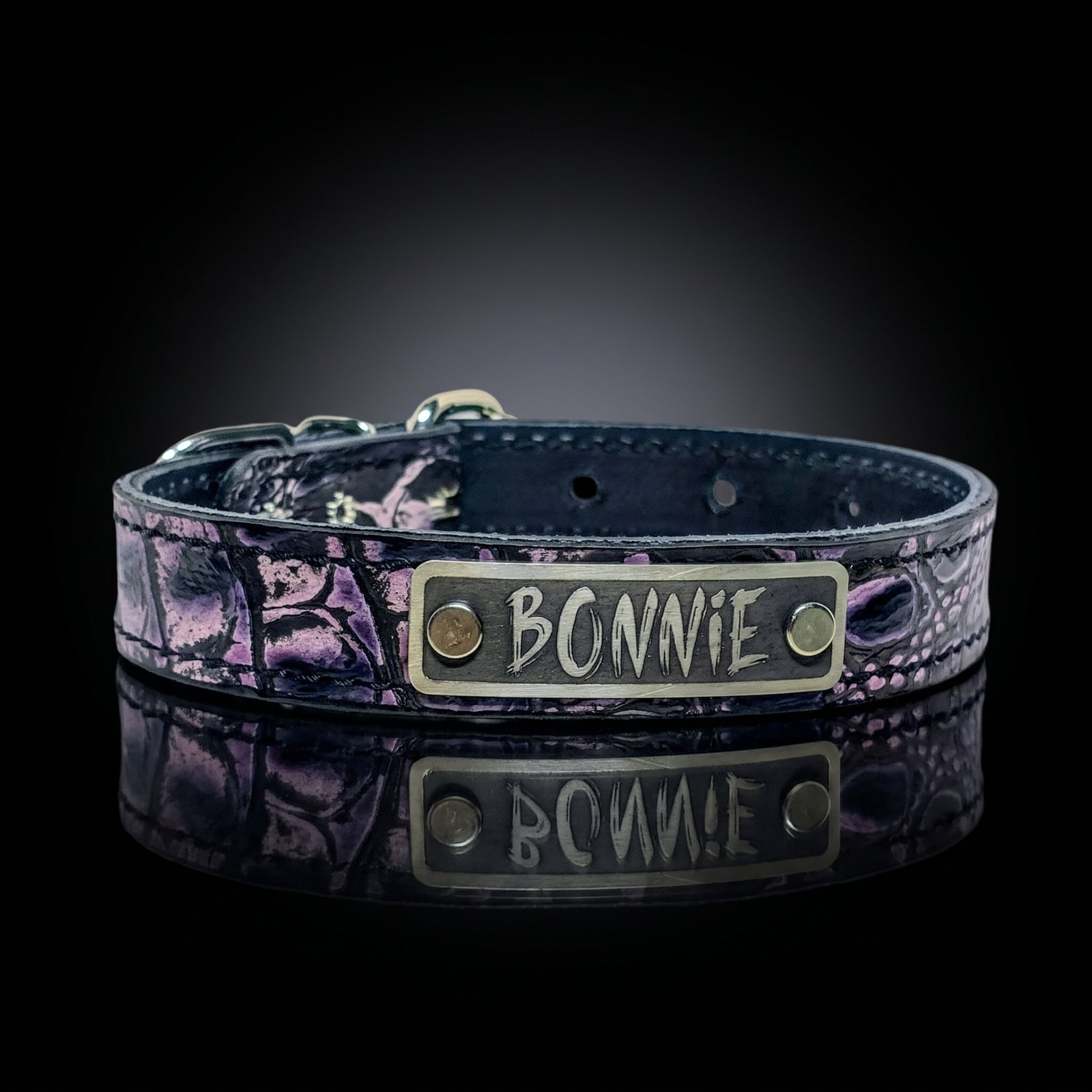 A purple leather collar with a purple and black textured pattern, featuring a brass nameplate engraved with 'BONNIE' and resting on a reflective black surface.