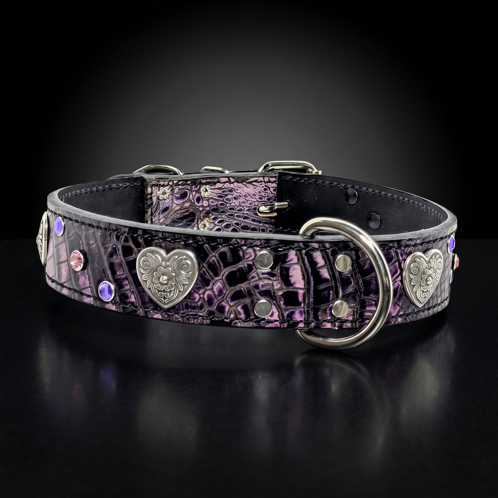 A decorative dog collar with a purple and black crocodile-embossed pattern, featuring silver-tone metal heart-shaped conchos, rhinestones, and a large D-ring, set against a dark reflective background.