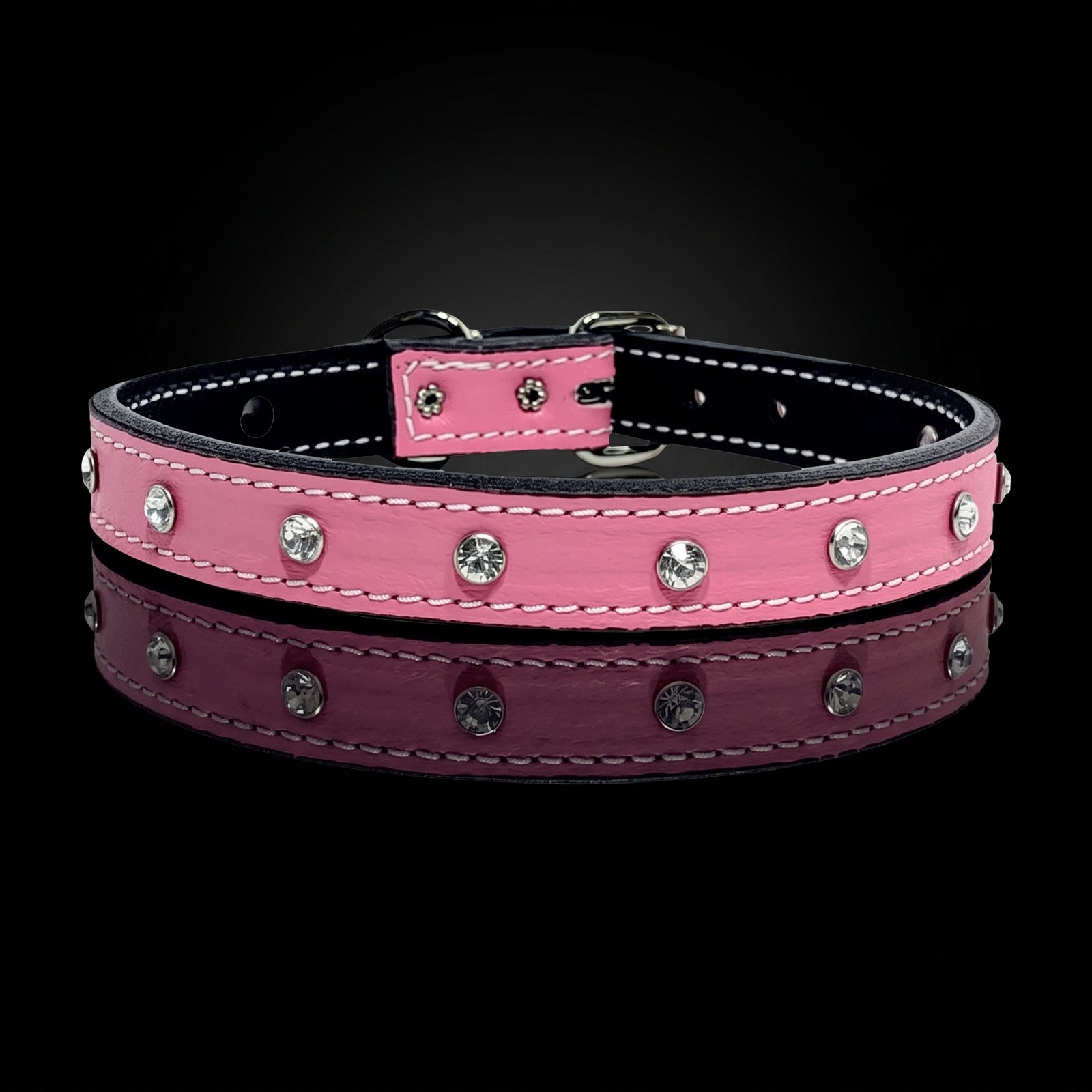 A pink leather dog collar with silver rhinestone studs and white contrast stitching, displayed on a reflective black surface against a black background.