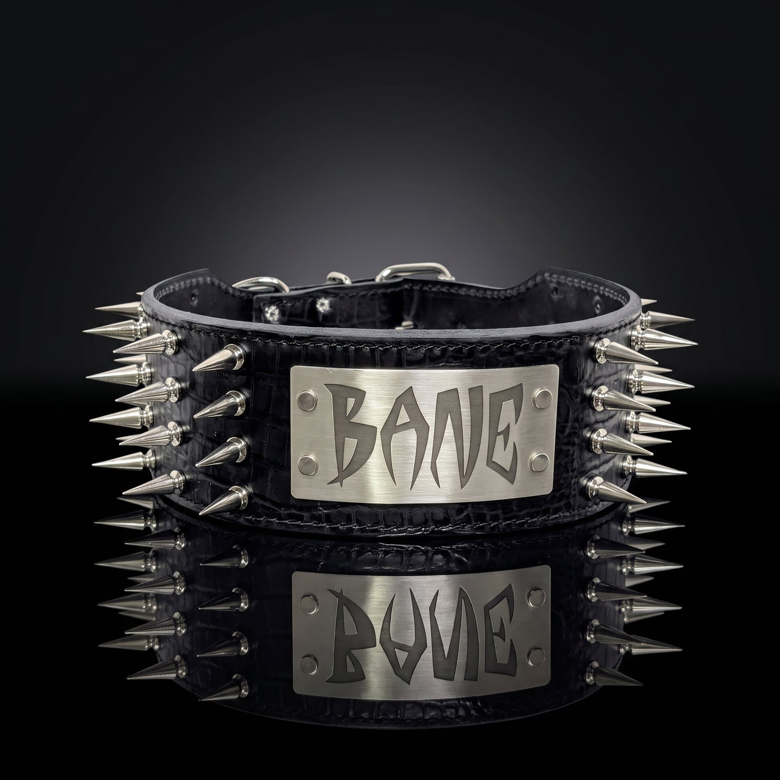 A black leather spiked collar with a silver metal plate engraved with the word 'BONE' in a stylized gothic font, displayed on a reflective black surface against a dark background.