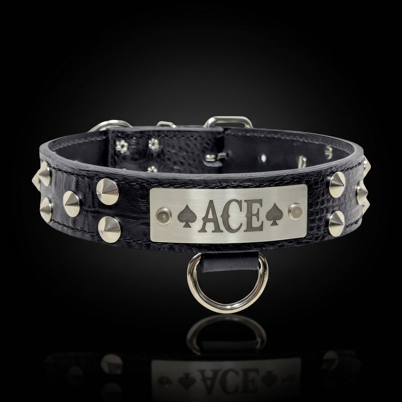 A black crocodile-embossed leather collar with silver-tone studs and a central silver metal plate engraved with 'ACE' and playing card suits, shown on a reflective black background.