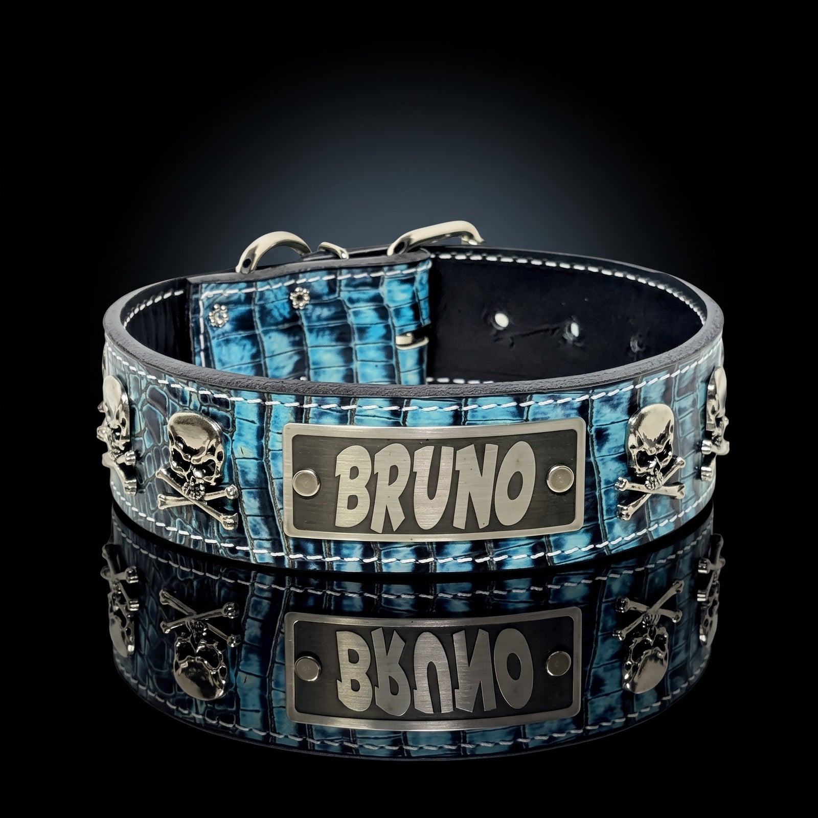 A blue and black crocodile-embossed leather pet collar with silver-tone skull and crossbones hardware and a central metal plaque engraved with the name 'BRUNO'. The collar is displayed on a reflective black surface against a dark gradient background.