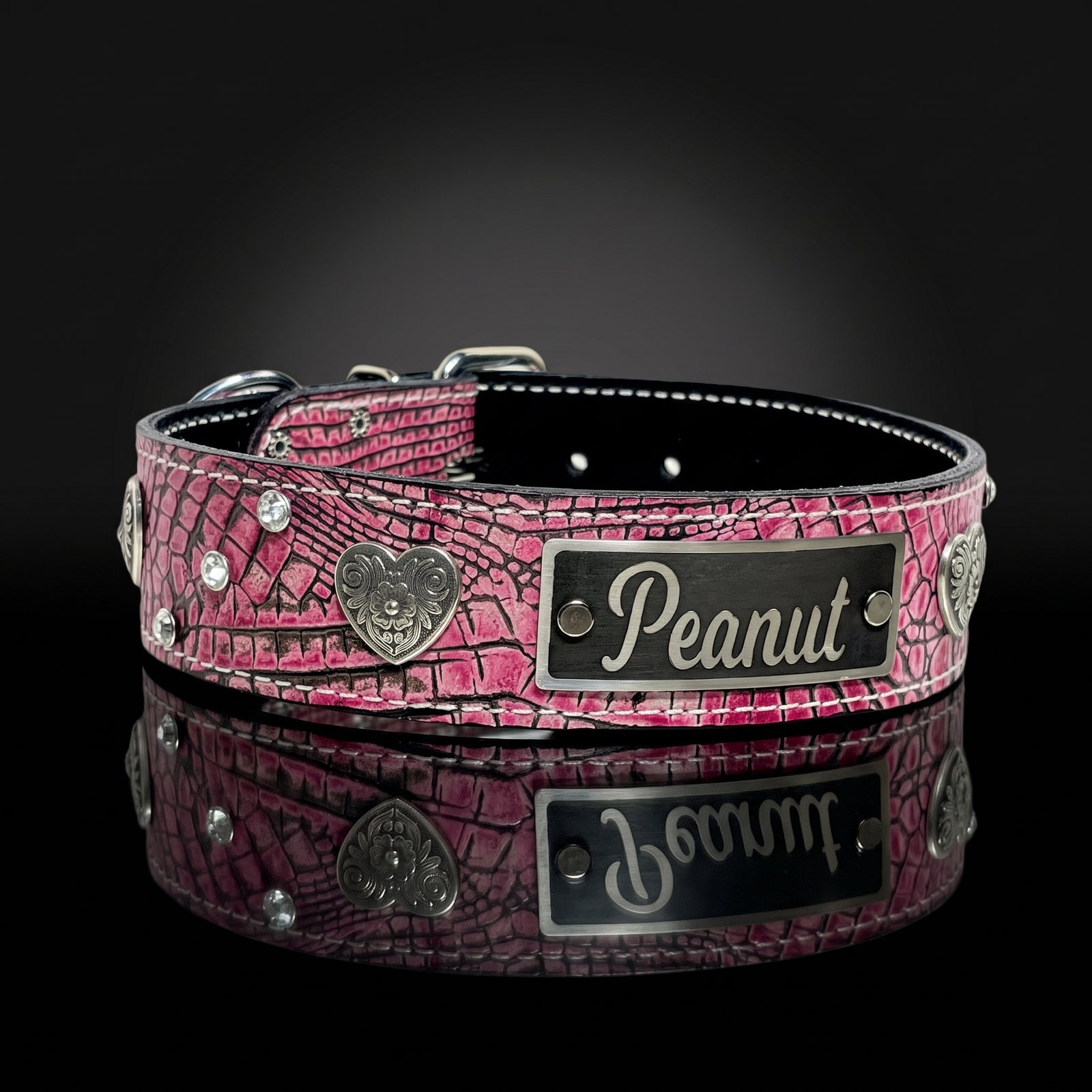 A pink crocodile-embossed leather dog collar with silver-tone metal hardware, including a heart-shaped emblem and a custom nameplate engraved with the name 'Peanut'. The collar is displayed on a reflective black surface against a dark background.