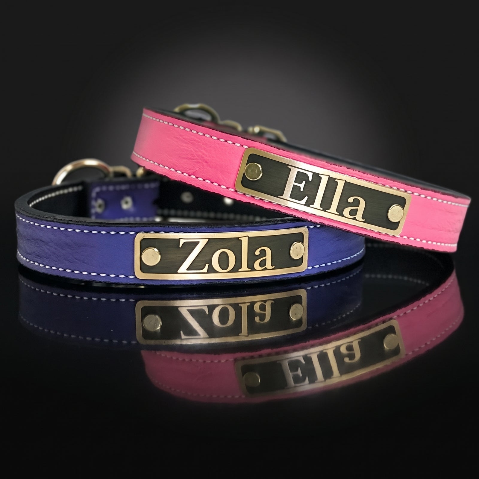 Two personalized dog collars, one pink and one blue, with gold metal nameplates reading 'Ella' and 'Zola' on a reflective black surface.