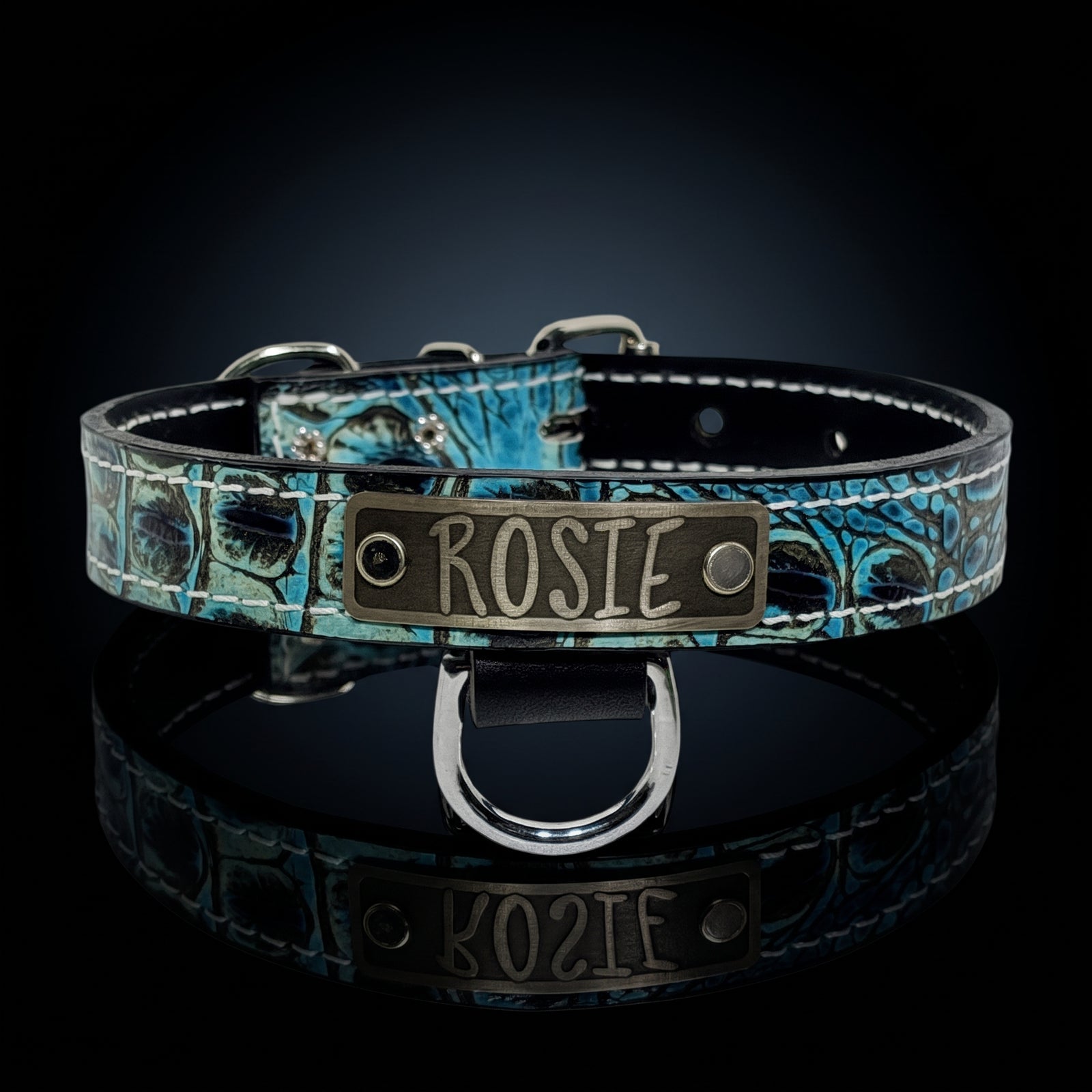 A personalized dog collar with a blue and black reptile pattern, featuring a silver metal buckle, D-ring, and a rectangular metal nameplate engraved with the name 'ROSIE'. The collar is displayed on a reflective black surface against a dark background.