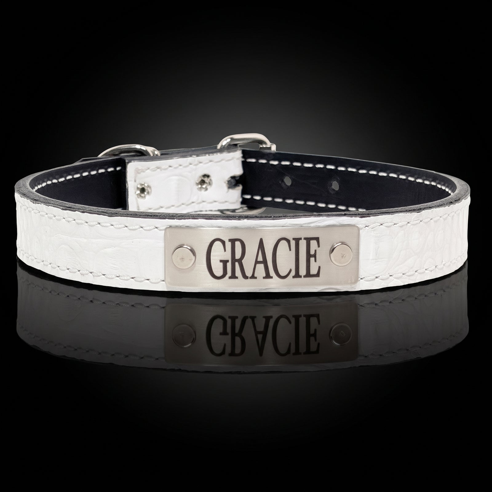 A personalized dog collar with a white textured faux-croc exterior, black interior, and a silver metal nameplate engraved with 'GRACIE'. The collar is displayed on a reflective black surface against a dark background.