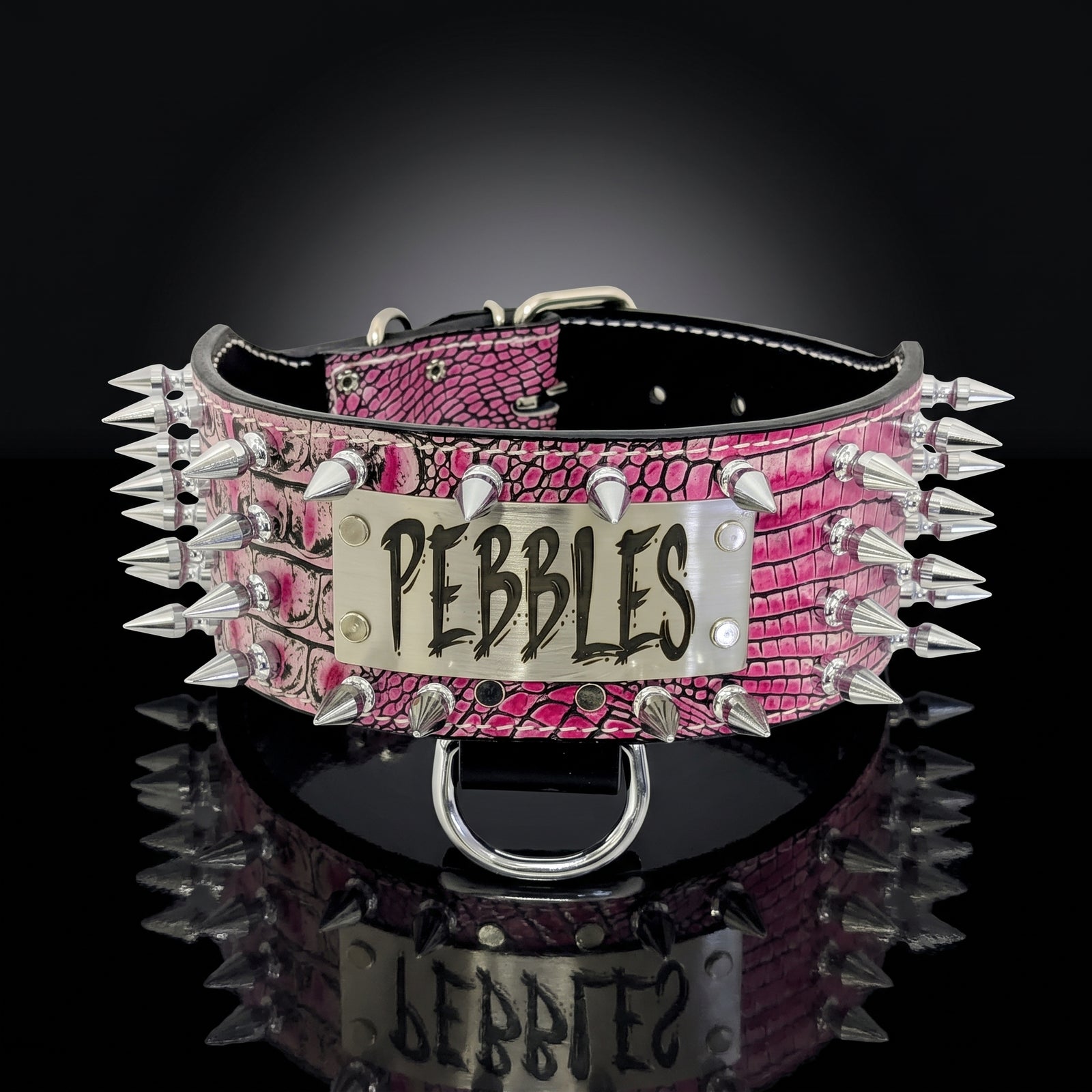 A pink crocodile-embossed spiked collar with a silver metal nameplate engraved 'PEBBLES' in black lettering, displayed on a reflective black surface against a dark background.