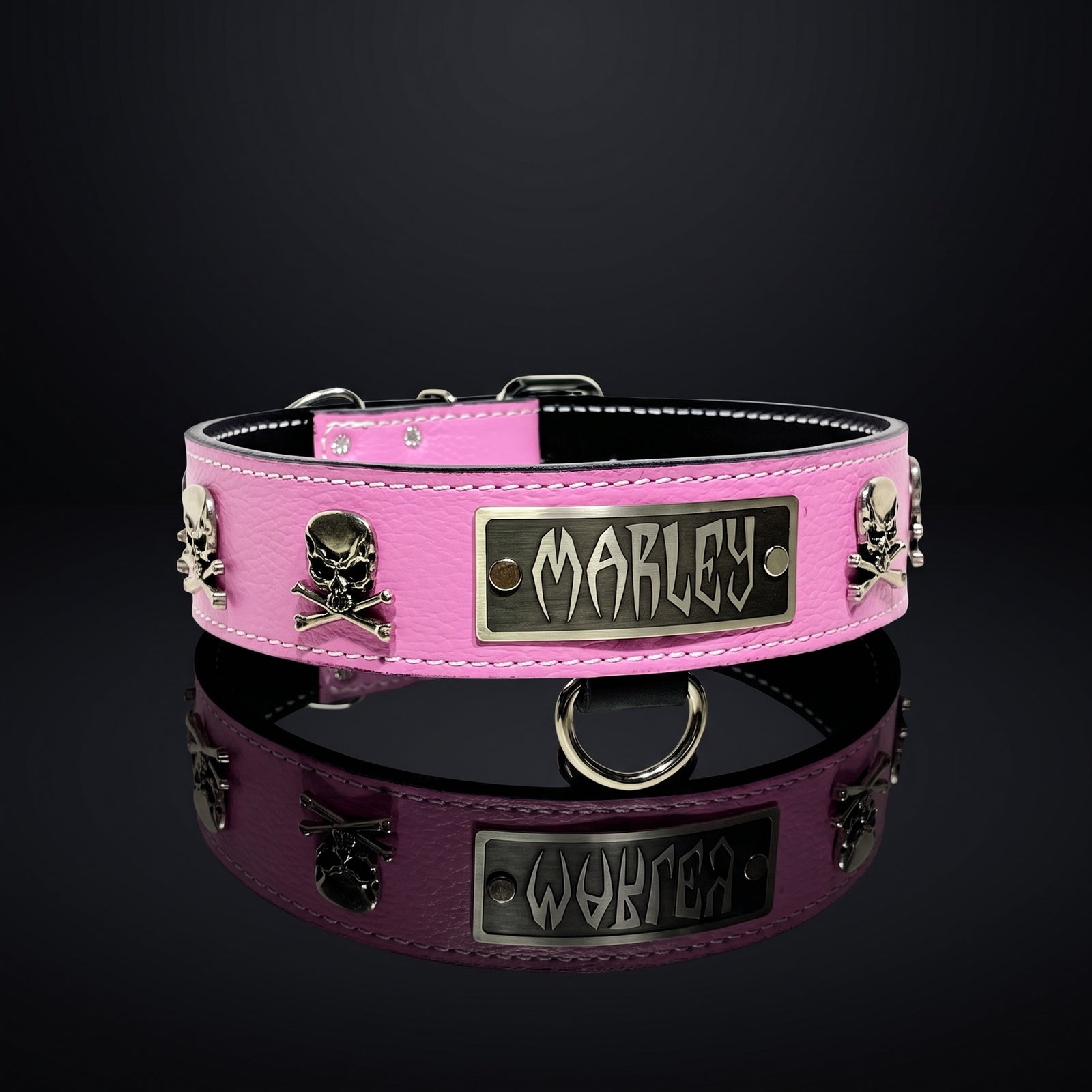 A pink leather dog collar with silver-tone skull and crossbones studs and a central rectangular plate engraved with the name 'MARLEY', shown on a reflective black surface against a dark background.