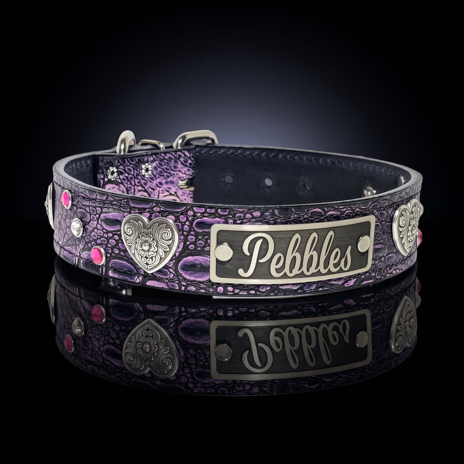 A custom pet collar with a purple and black crocodile-embossed pattern, featuring silver metal accents, a heart-shaped skull emblem, and a rectangular nameplate engraved with the name 'Pebbles'. The collar is displayed on a reflective black surface.