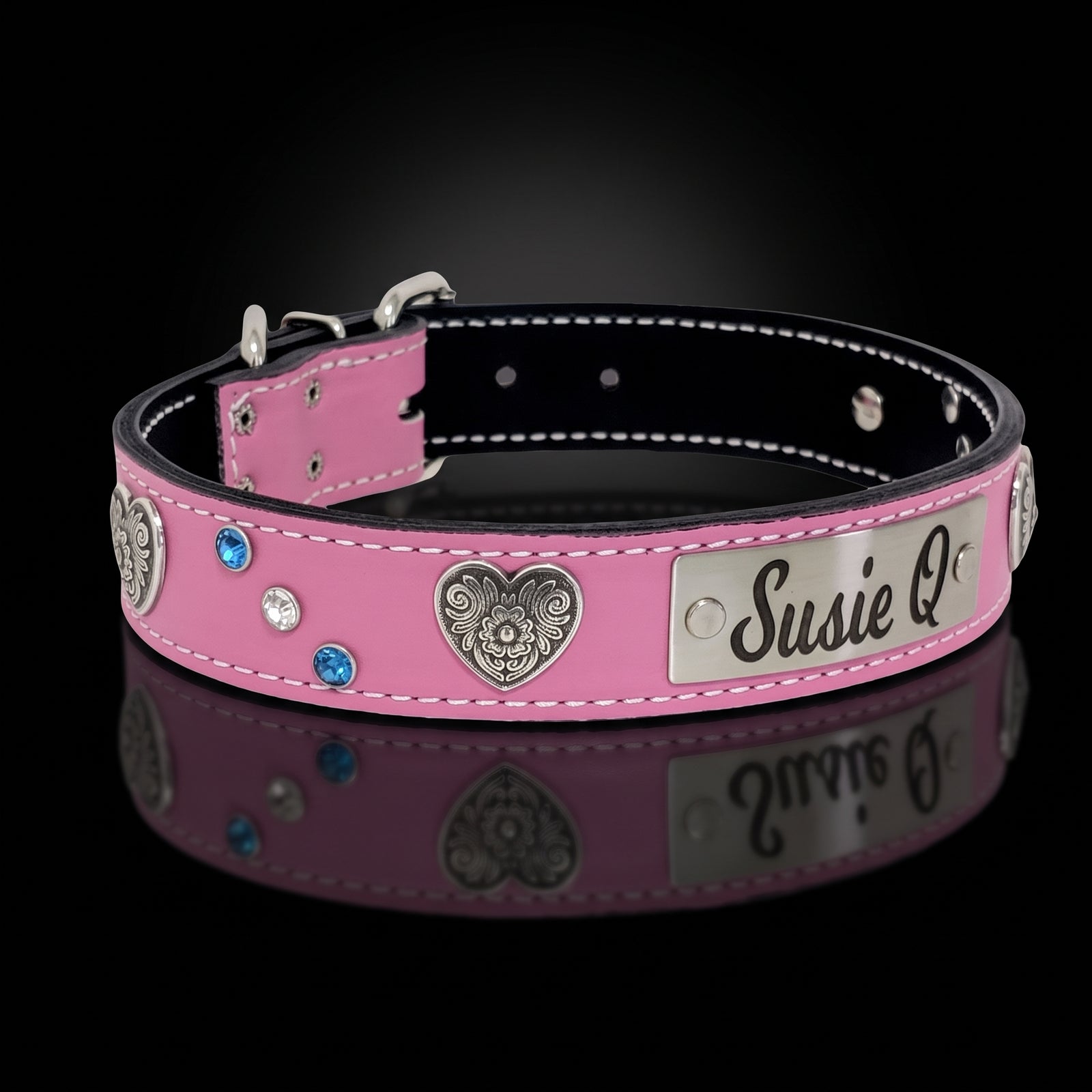 A pink leather dog collar with silver-tone metal hardware, featuring a heart-shaped filigree ornament, blue and clear rhinestones, and a custom engraved silver nameplate that reads 'Susie Q'. The collar is displayed on a reflective black surface against a dark gradient background.