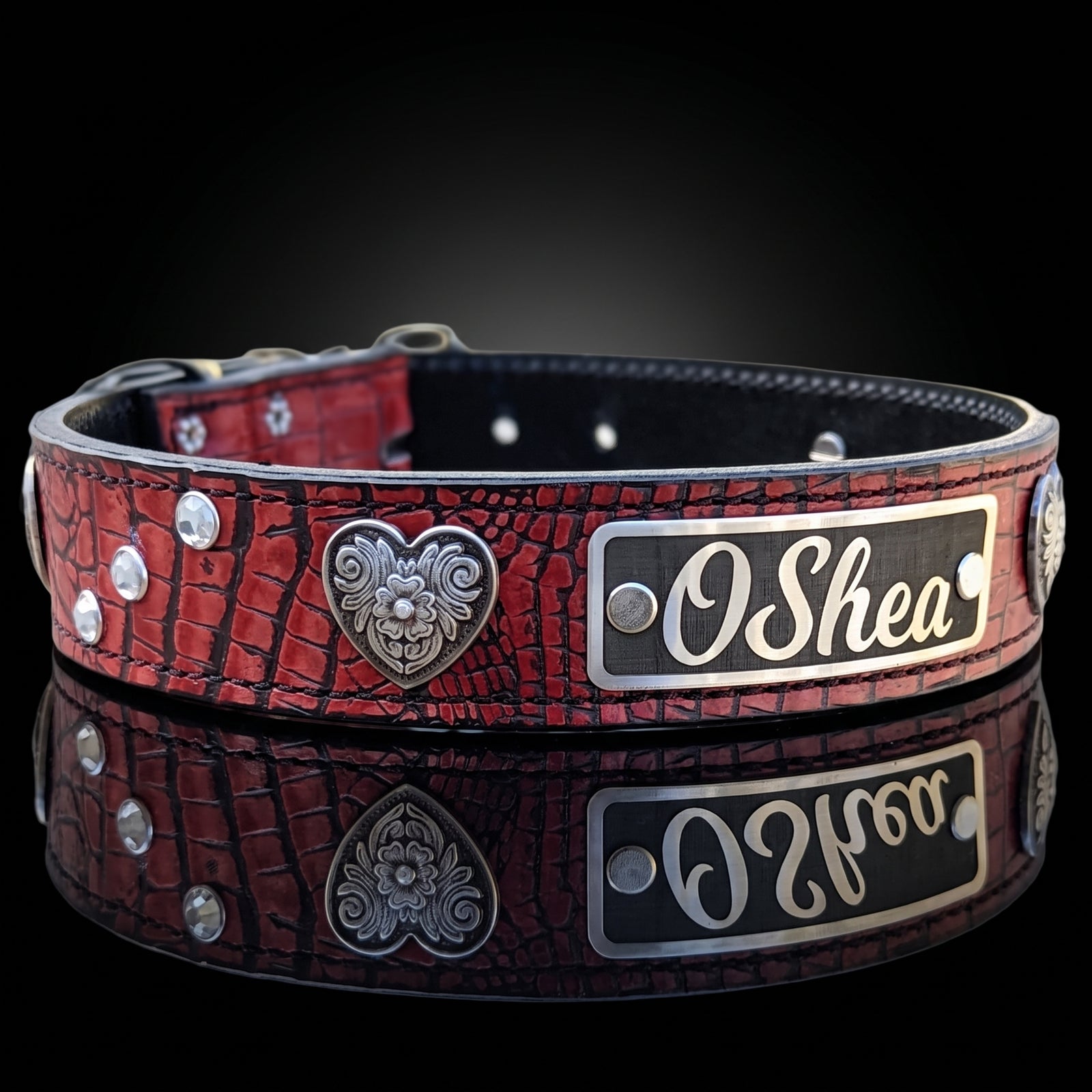 A red crocodile-embossed leather dog collar with silver-tone hardware, featuring a heart-shaped filigree emblem and a rectangular nameplate engraved with 'O Shea'. The collar is displayed on a reflective black surface against a dark background.