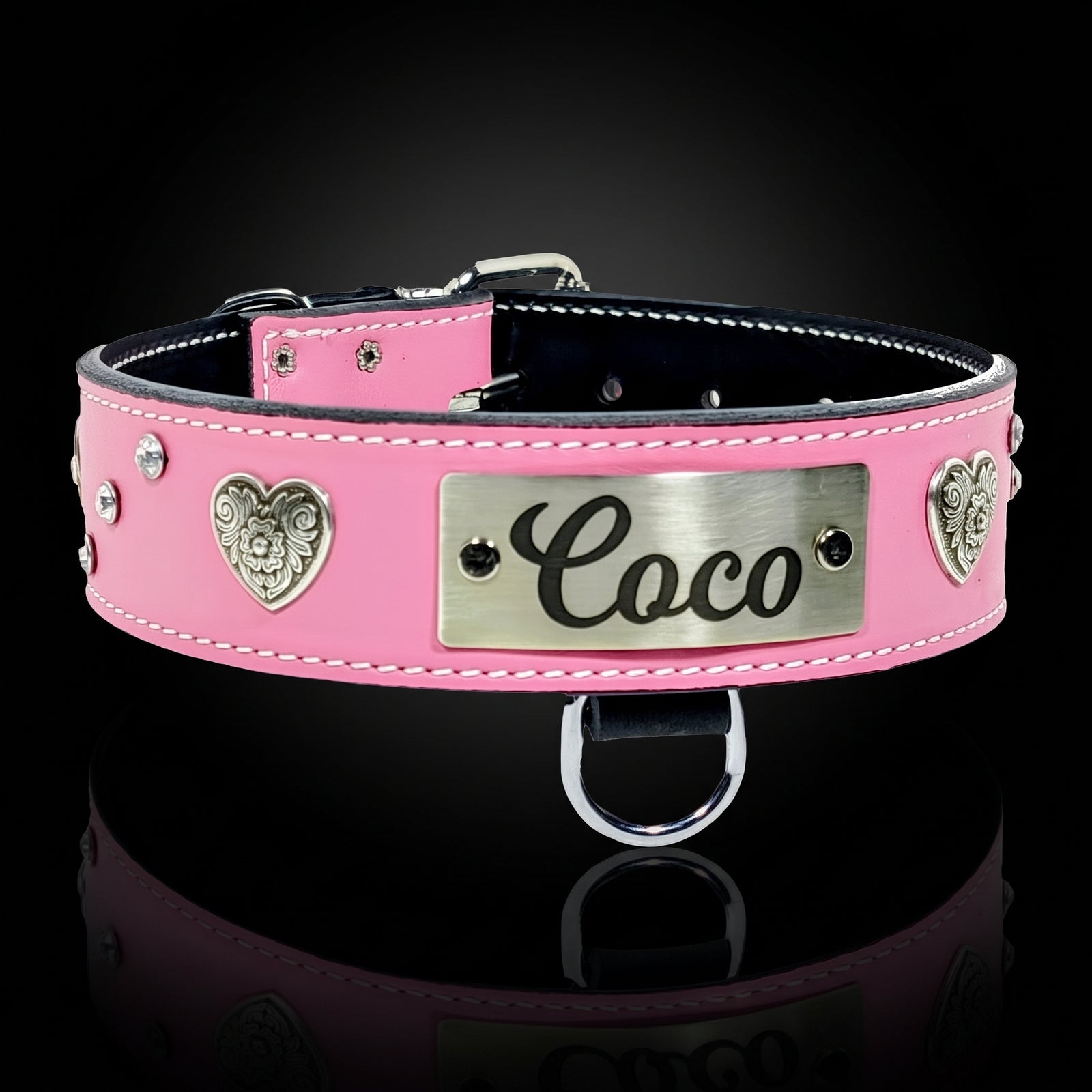 A pink leather dog collar with silver-tone metal hardware, featuring heart-shaped studs, a central rectangular plate engraved with the name 'Coco', and a hanging ring, shown on a black reflective background.