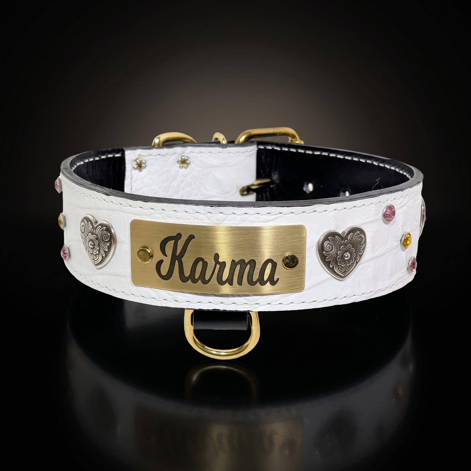 A white crocodile-embossed leather dog collar with a brushed gold metal nameplate engraved with 'Karma', flanked by decorative silver heart charms and small colored rhinestones, shown on a reflective black background.