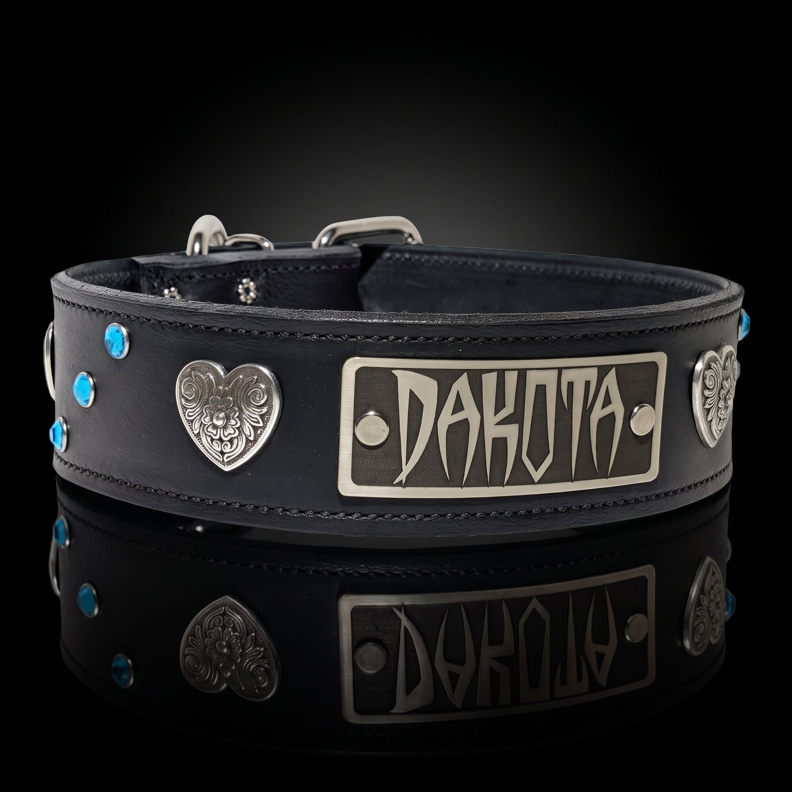 A black leather collar with silver-tone metal hardware, featuring a central rectangular plate engraved with 'DAKOTA', heart-shaped medallions, and small blue rhinestone accents, displayed on a reflective black surface.