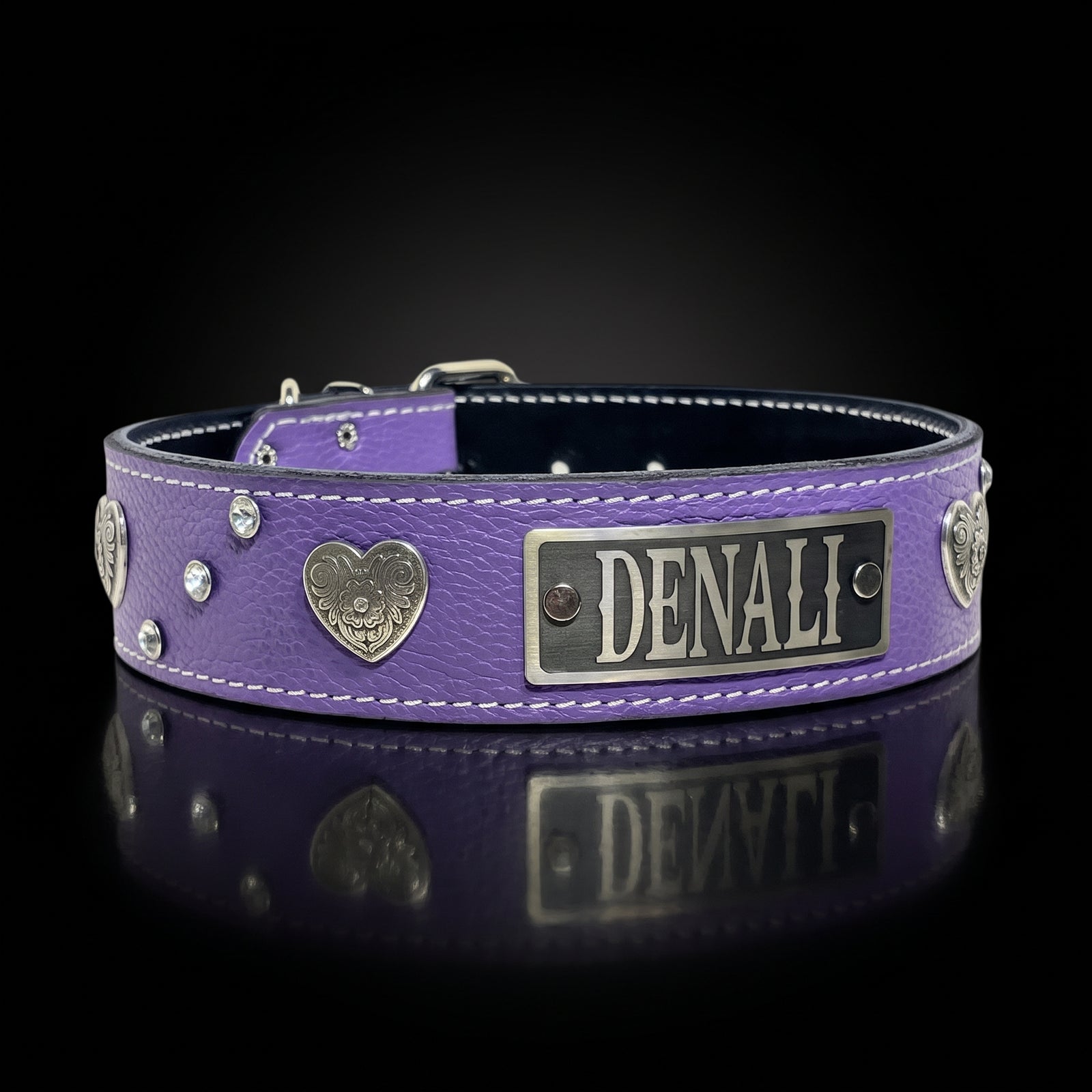 A purple leather dog collar with silver-tone hardware, including a rectangular nameplate engraved 'DENALI', heart-shaped medallions, and rhinestone accents, displayed on a reflective black surface.