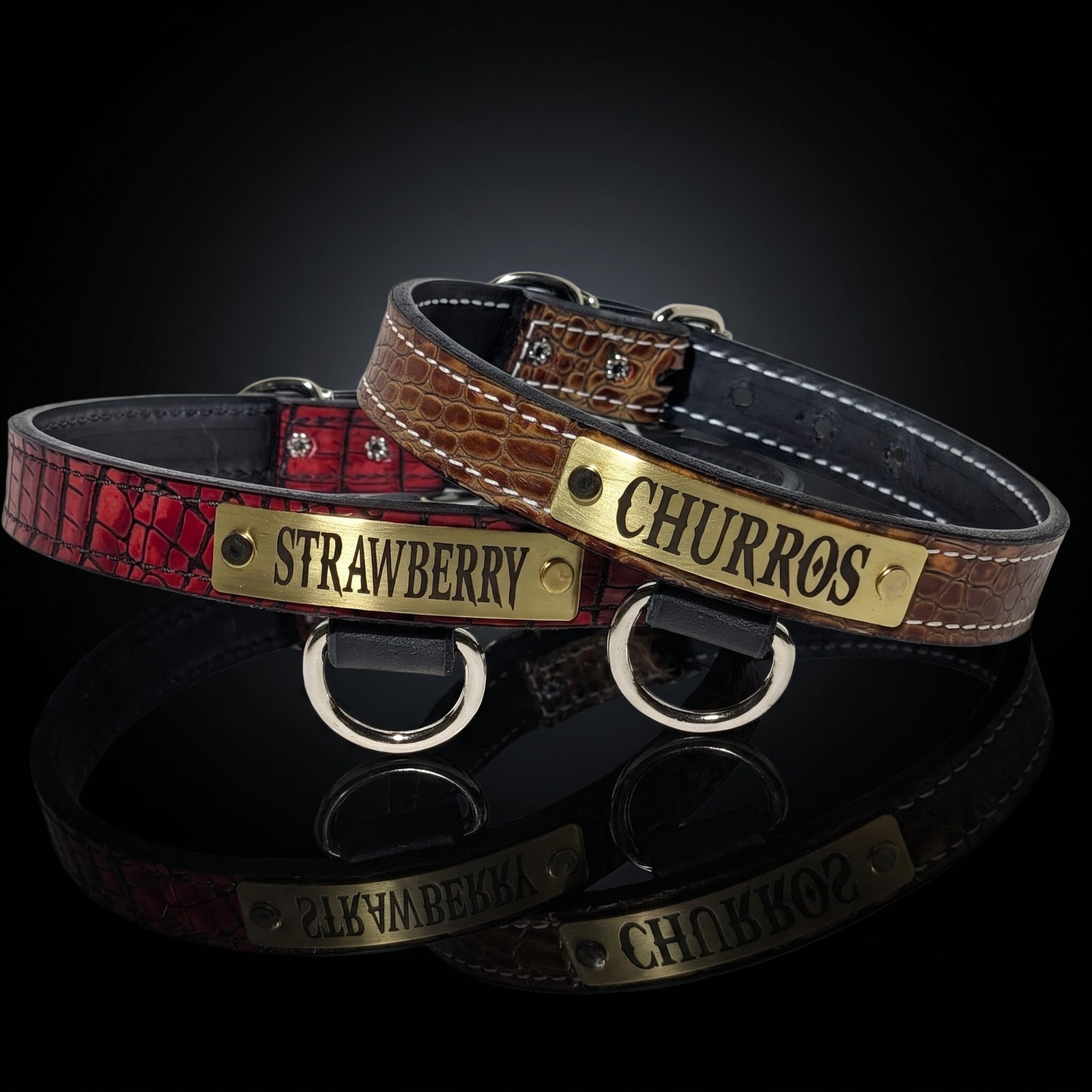 Two personalized dog collars displayed on a reflective black surface. One collar is textured red with a brass plate engraved 'STRAWBERRY', and the other is brown crocodile-embossed with a brass plate engraved 'CHURROS'.