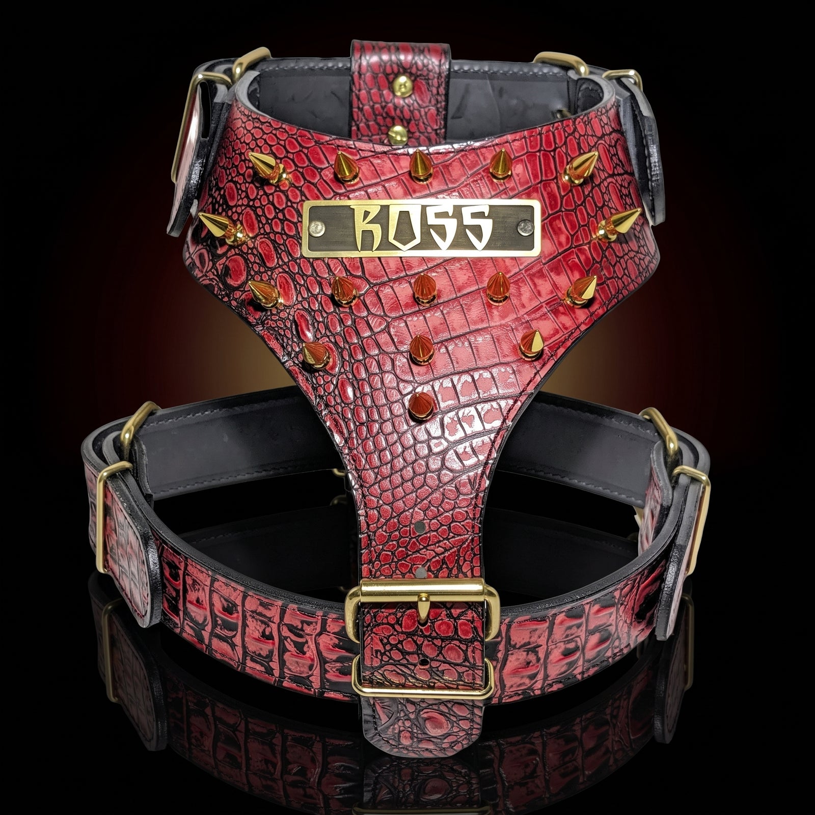 A red crocodile-embossed leather dog harness with gold-tone hardware, featuring a central brass plate engraved with 'BOSS' and gold spikes along the sides, set against a dark reflective background.
