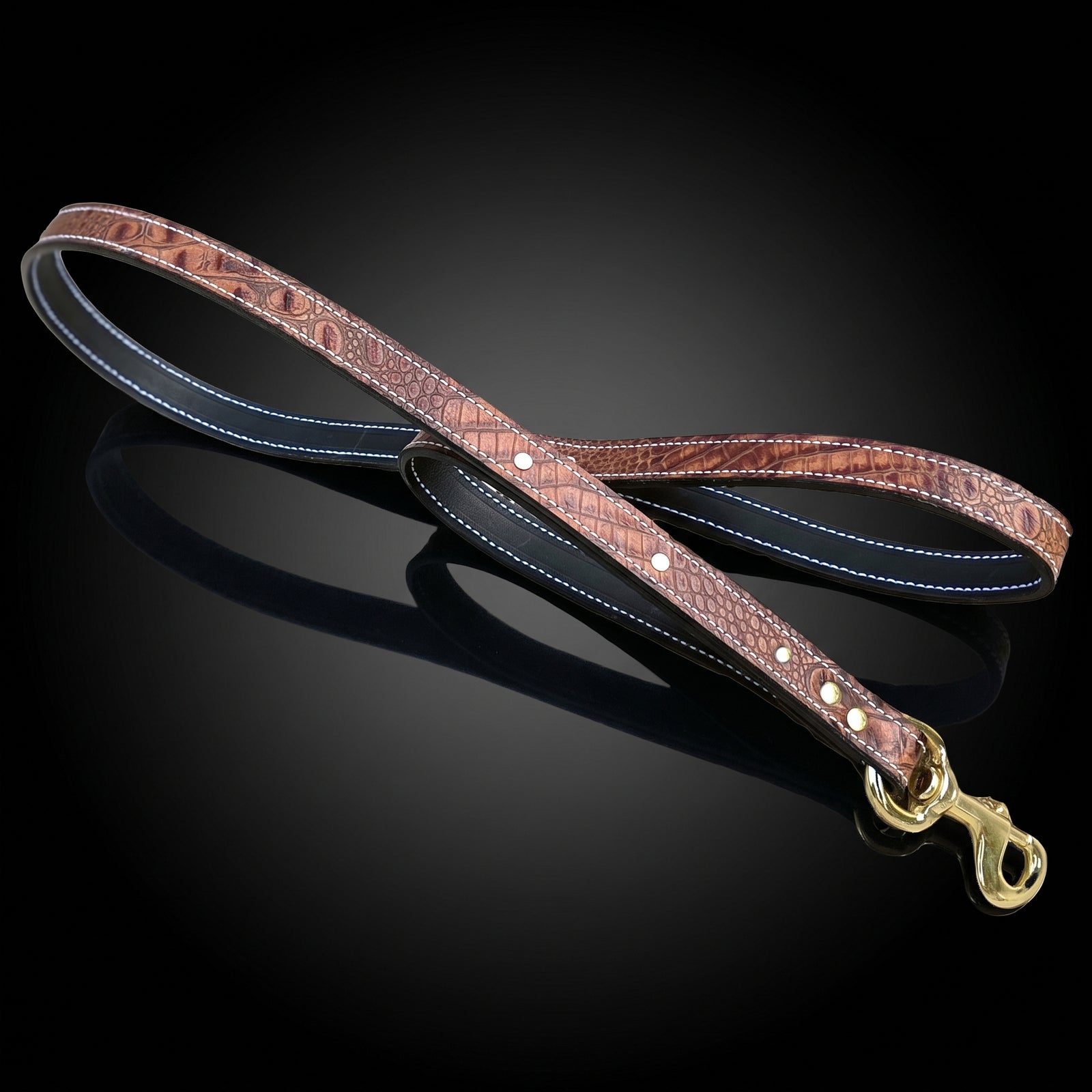 A brown crocodile-embossed leather dual-handle dog leash with white contrast stitching and a brass snap hook, coiled on a dark, reflective wet obsidian surface.