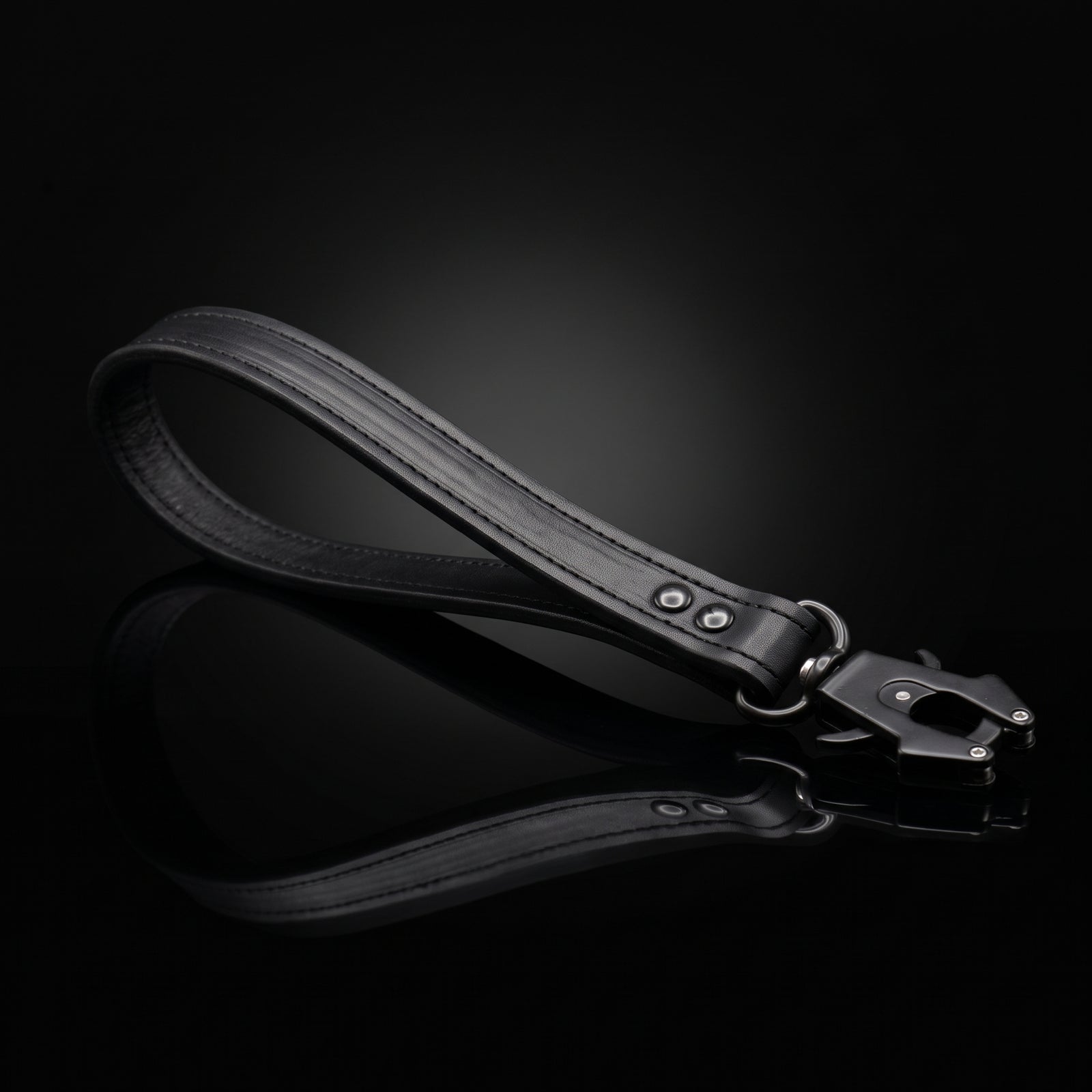 A black leather strap accessory with metal hardware, photographed on a glossy black reflective surface against a dark studio background.