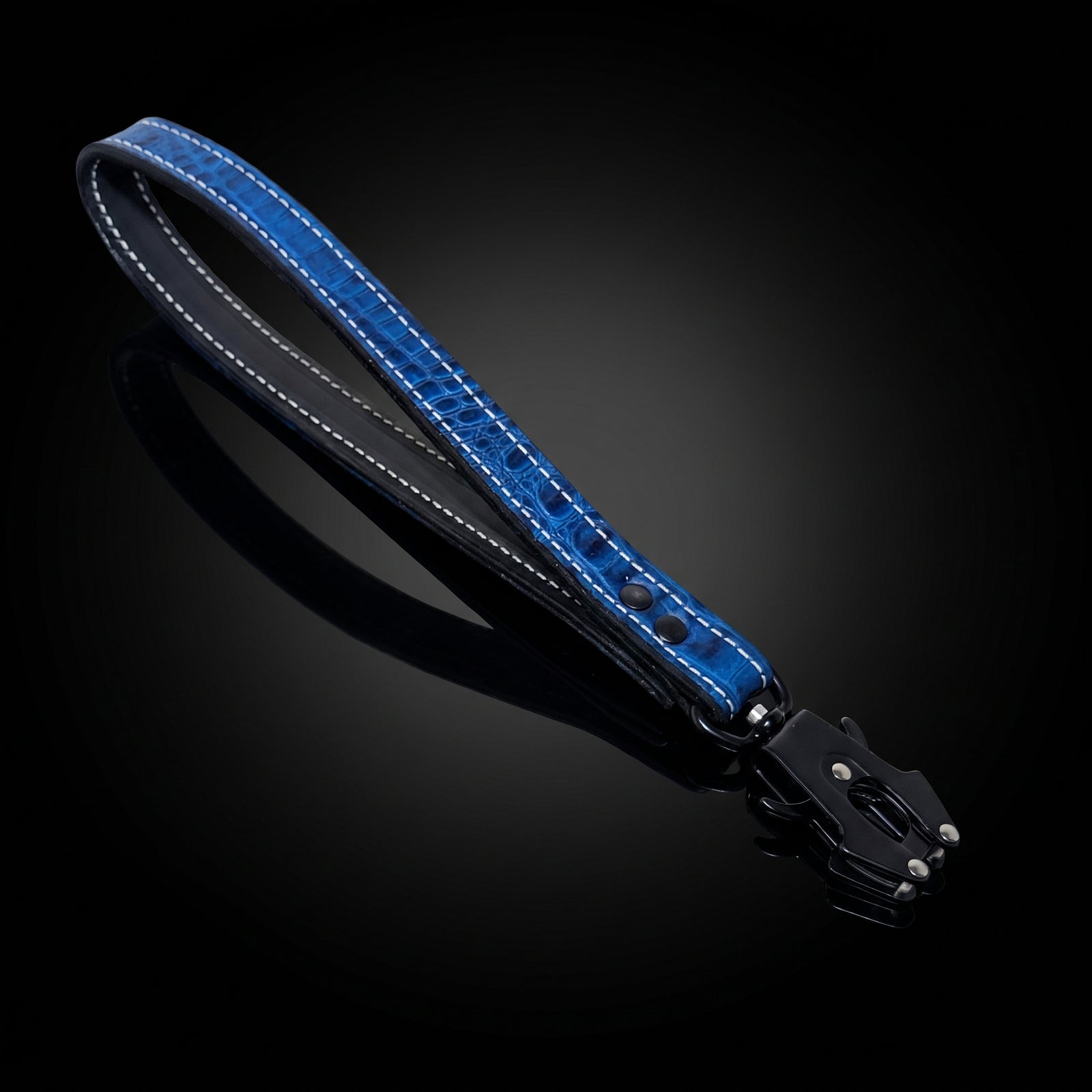A blue crocodile-embossed leather strap with white contrast stitching and a black metal clasp, displayed diagonally on a reflective black background.