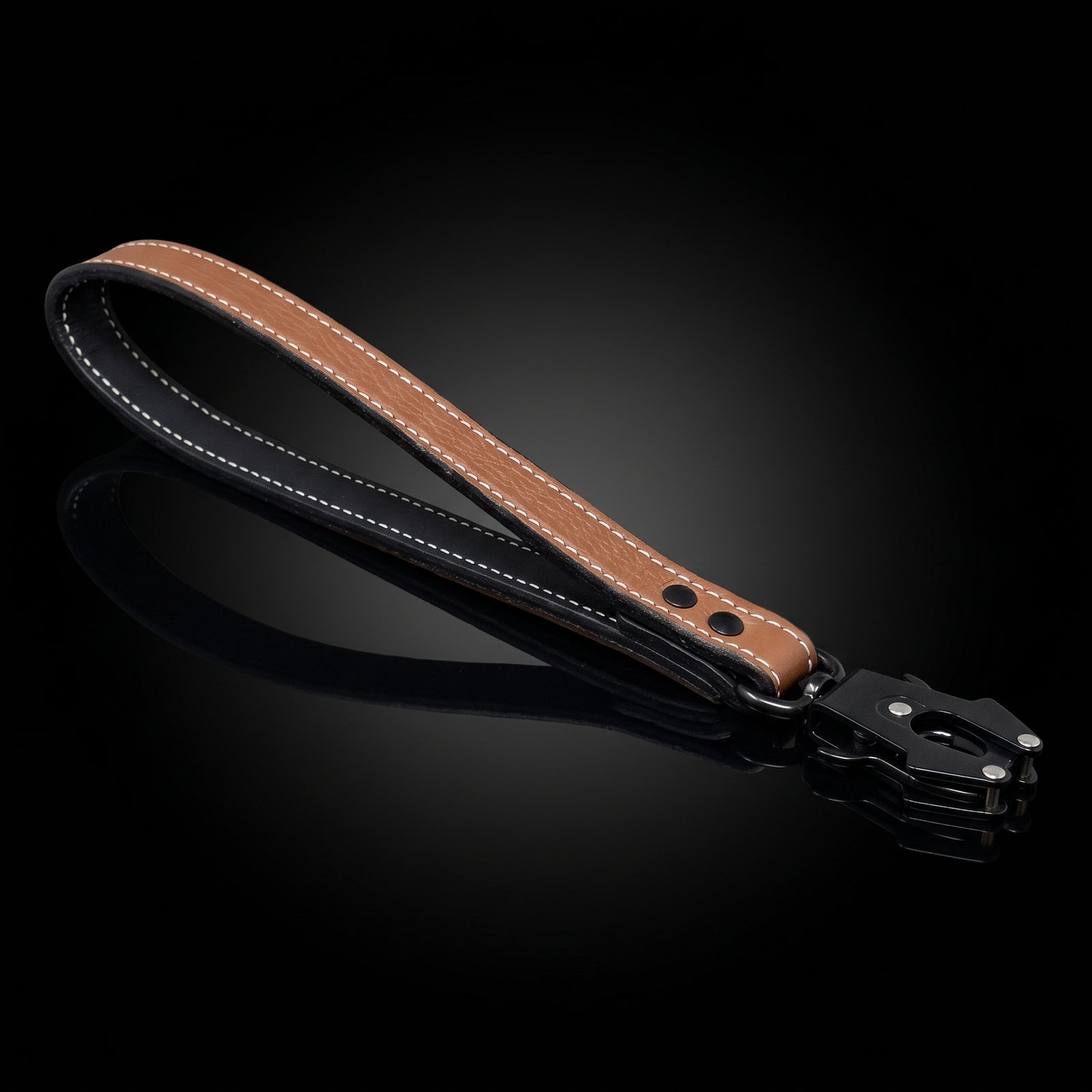 A brown leather wrist strap with white contrast stitching and a black metal clasp, displayed on a reflective black background.