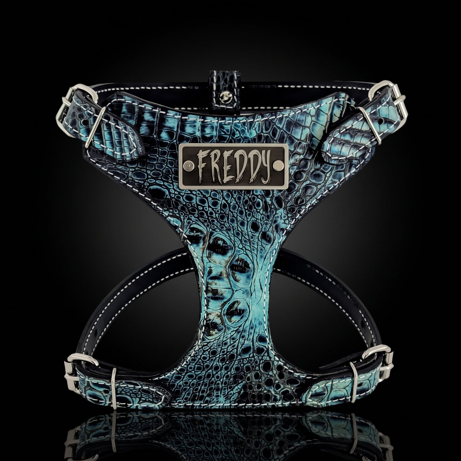 A dog harness with a textured turquoise and black crocodile pattern, featuring a central metal plate engraved with the name 'FREDDY'.