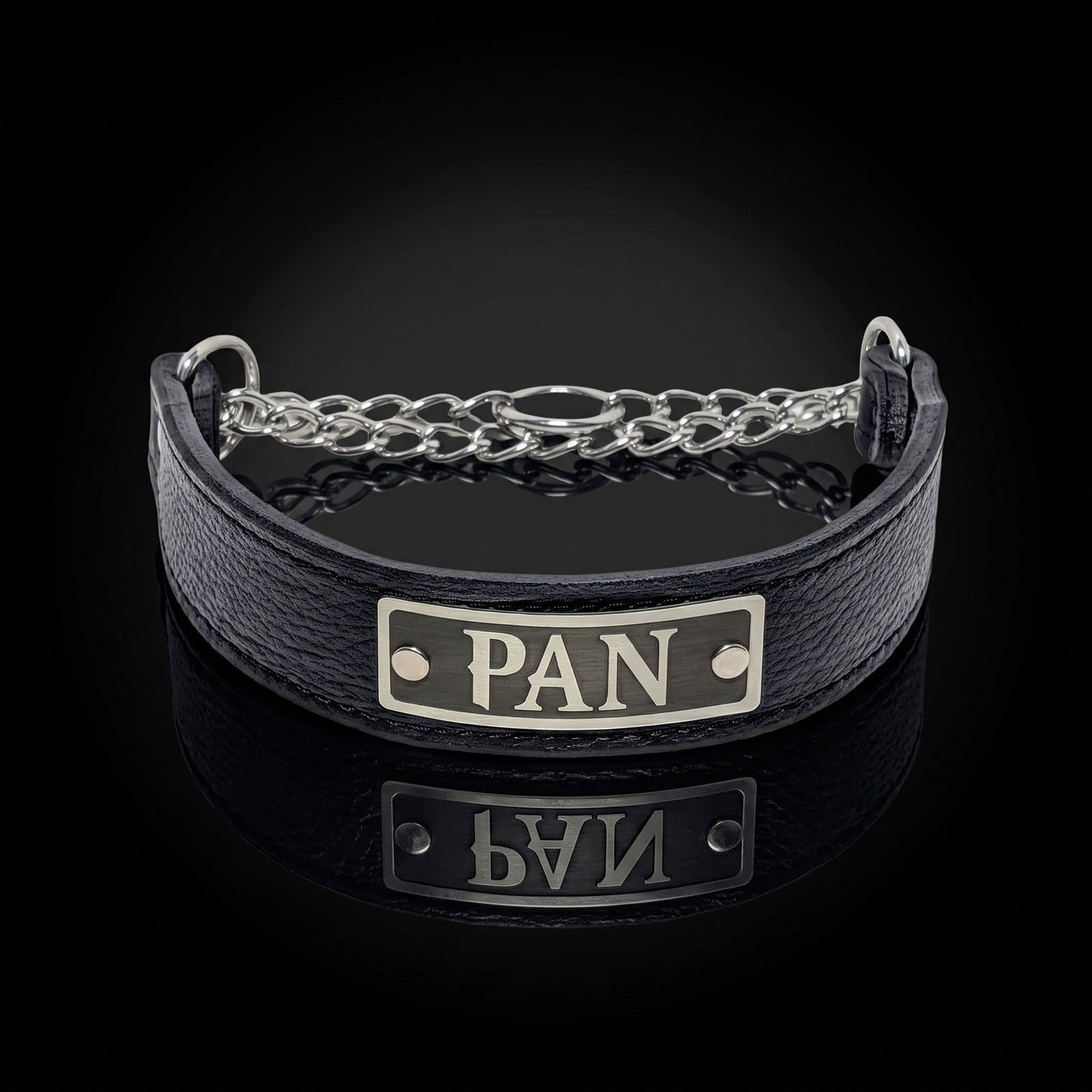 A black textured leather bracelet with a silver metal chain link accent and a rectangular silver plate engraved with the letters 'PAN', shown on a reflective black background.