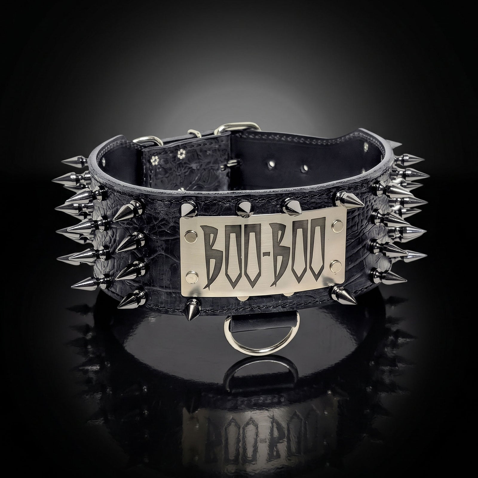 A black crocodile-embossed leather dog collar with silver spikes and a central silver metal plate engraved with 'BOO-BOO', shown on a reflective black background.