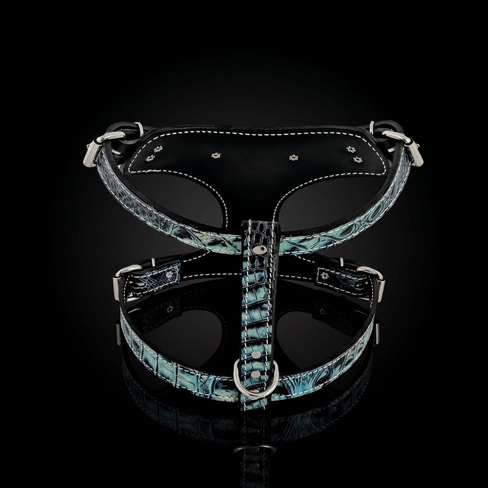 A black leather dog harness with a blue and green mottled reptile-textured pattern on the straps, featuring silver metal hardware and white contrast stitching, displayed on a reflective black background.