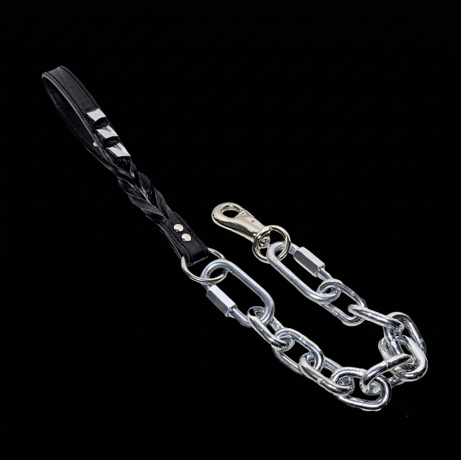 A super heavy silver chain dog leash featuring a black leather handle with twisted braided detailing and silver-tone decorative spikes, attached to a robust metal snap hook, displayed against a cinematic black background.
