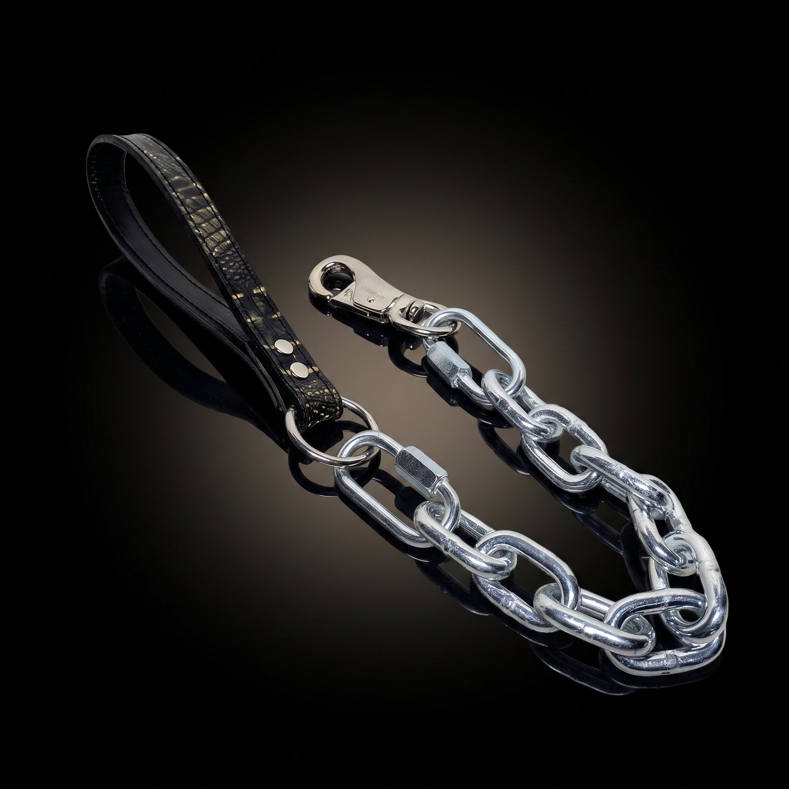 A black leather strap accessory with gold studs and a silver metal chain link connector, displayed on a reflective black background.
