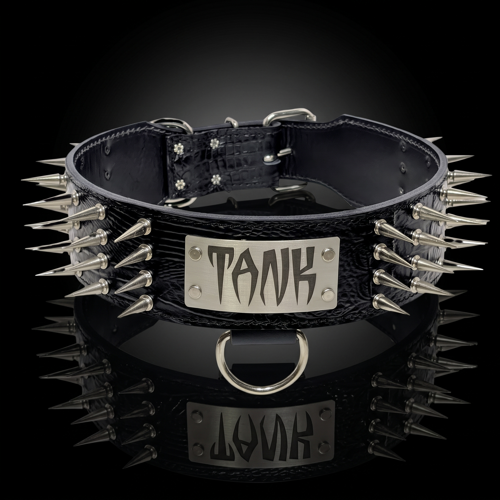 A black leather spiked collar with a silver metal plaque engraved with the word 'TANK' in a stylized font, displayed on a reflective black surface.