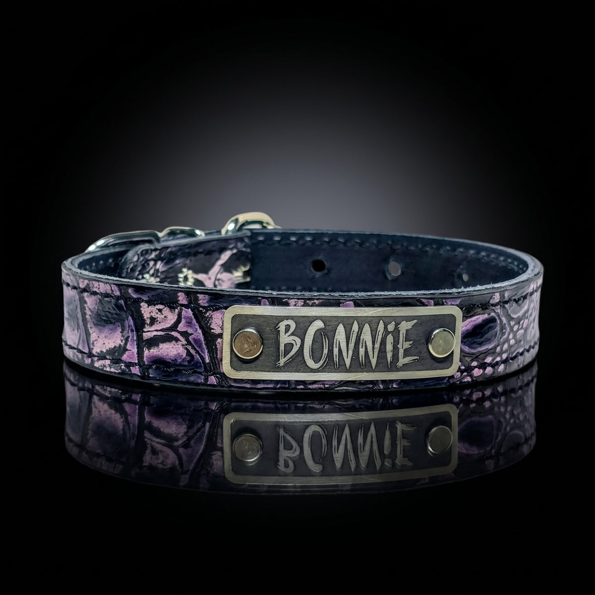 A purple leather collar with a purple and black textured pattern, featuring a brass nameplate engraved with 'BONNIE' and resting on a reflective black surface.