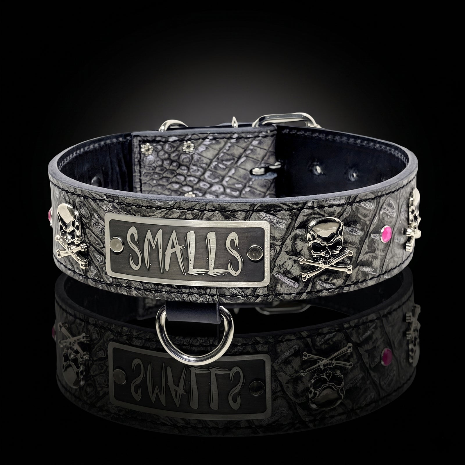A black textured leather dog collar with skull and crossbones details, a central silver-tone metal plate engraved with 'SMALLS', and pink rhinestone accents, displayed on a reflective black surface.