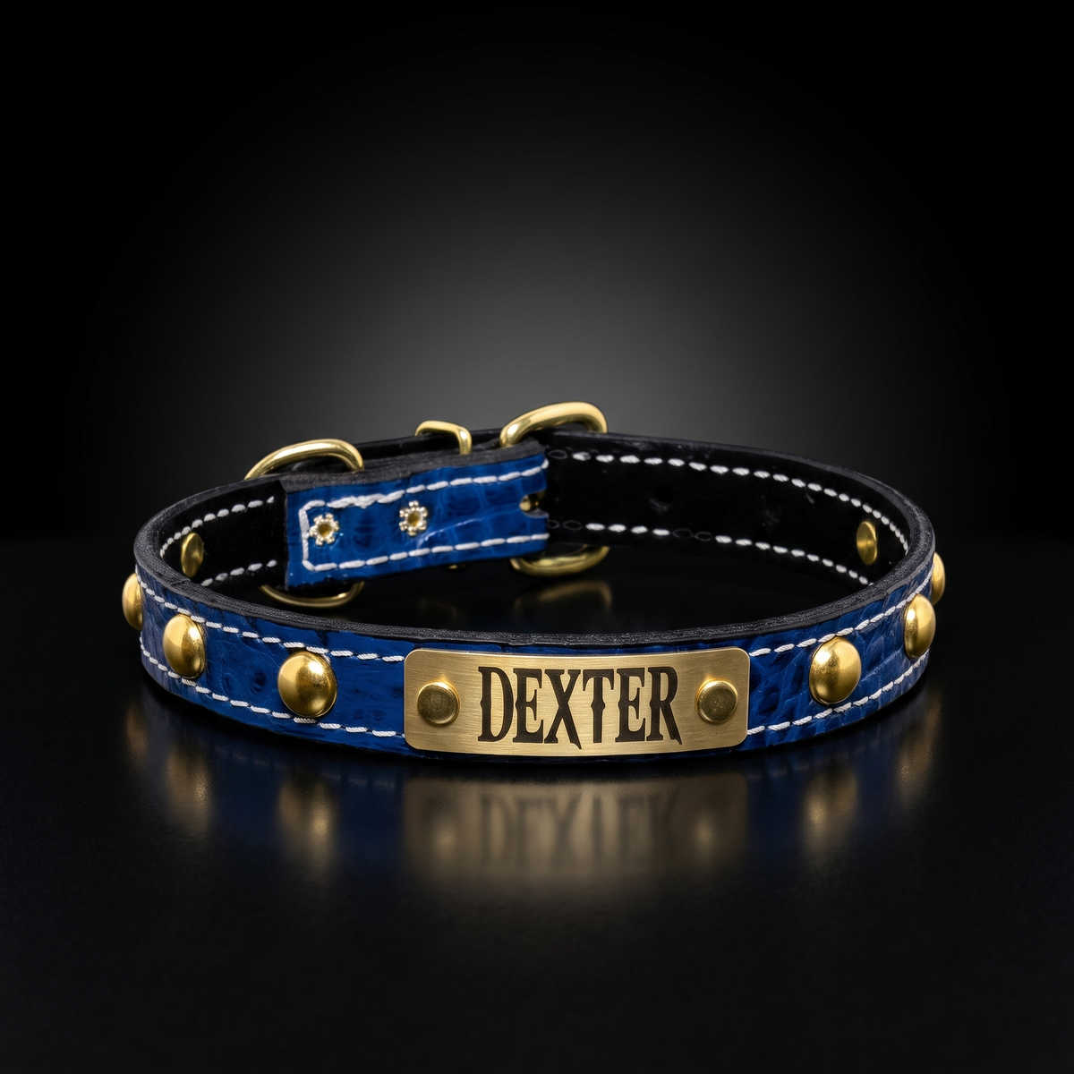 A personalized blue crocodile-embossed leather dog collar with gold-tone hardware, white stitching, and a brass nameplate engraved with the name 'DEXTER'.