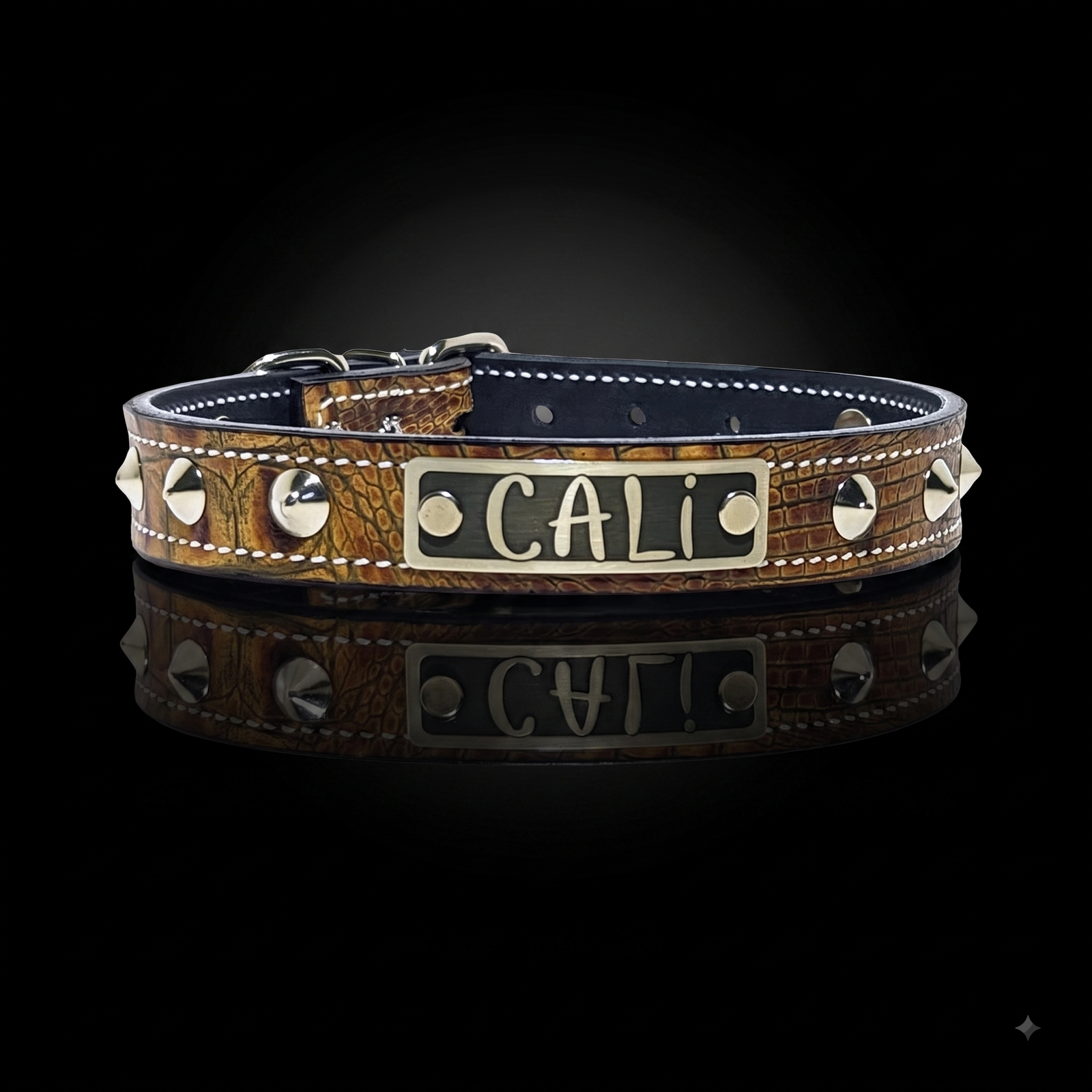 A brown crocodile-embossed leather dog collar with silver-tone hardware, white stitching, and a central metal plate engraved with the name 'Cali', displayed on a reflective black background.