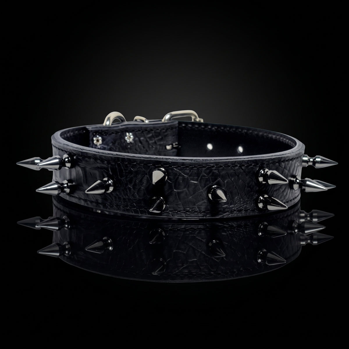 A black crocodile-embossed leather dog collar with sharp silver metal spikes and studs, displayed on a reflective black surface against a dark background.