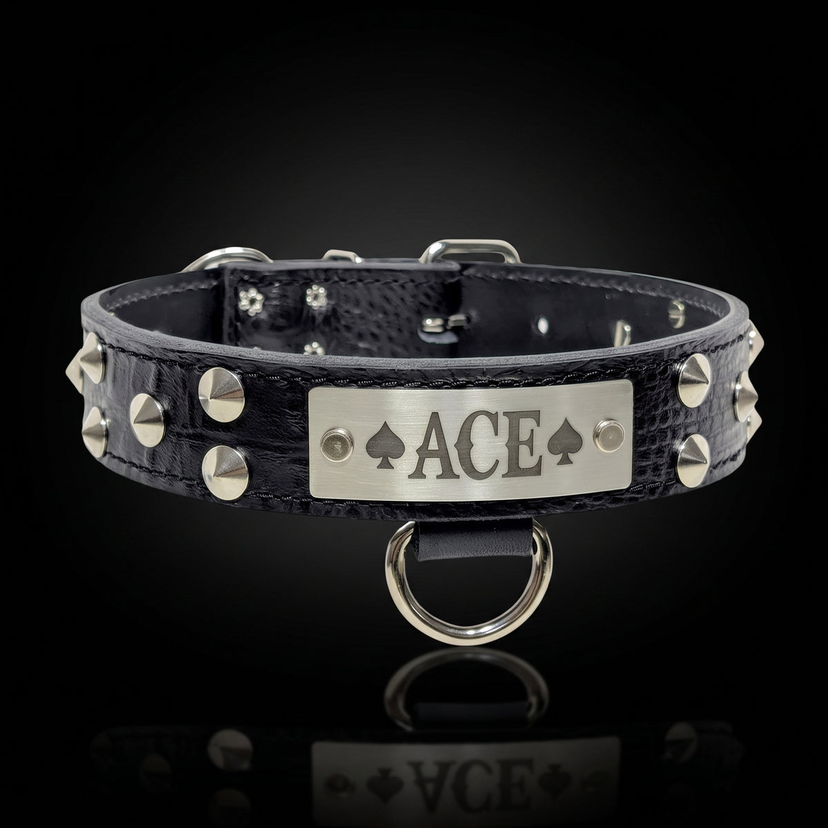 A black crocodile-embossed leather collar with silver-tone studs and a central silver metal plate engraved with 'ACE' and playing card suits, shown on a reflective black background.