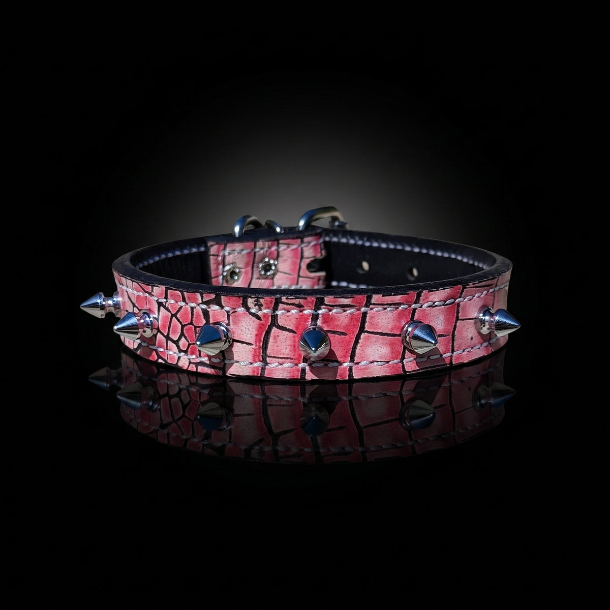 A pink crocodile-embossed leather collar with silver metal spikes and studs, displayed on a reflective black surface against a dark background.