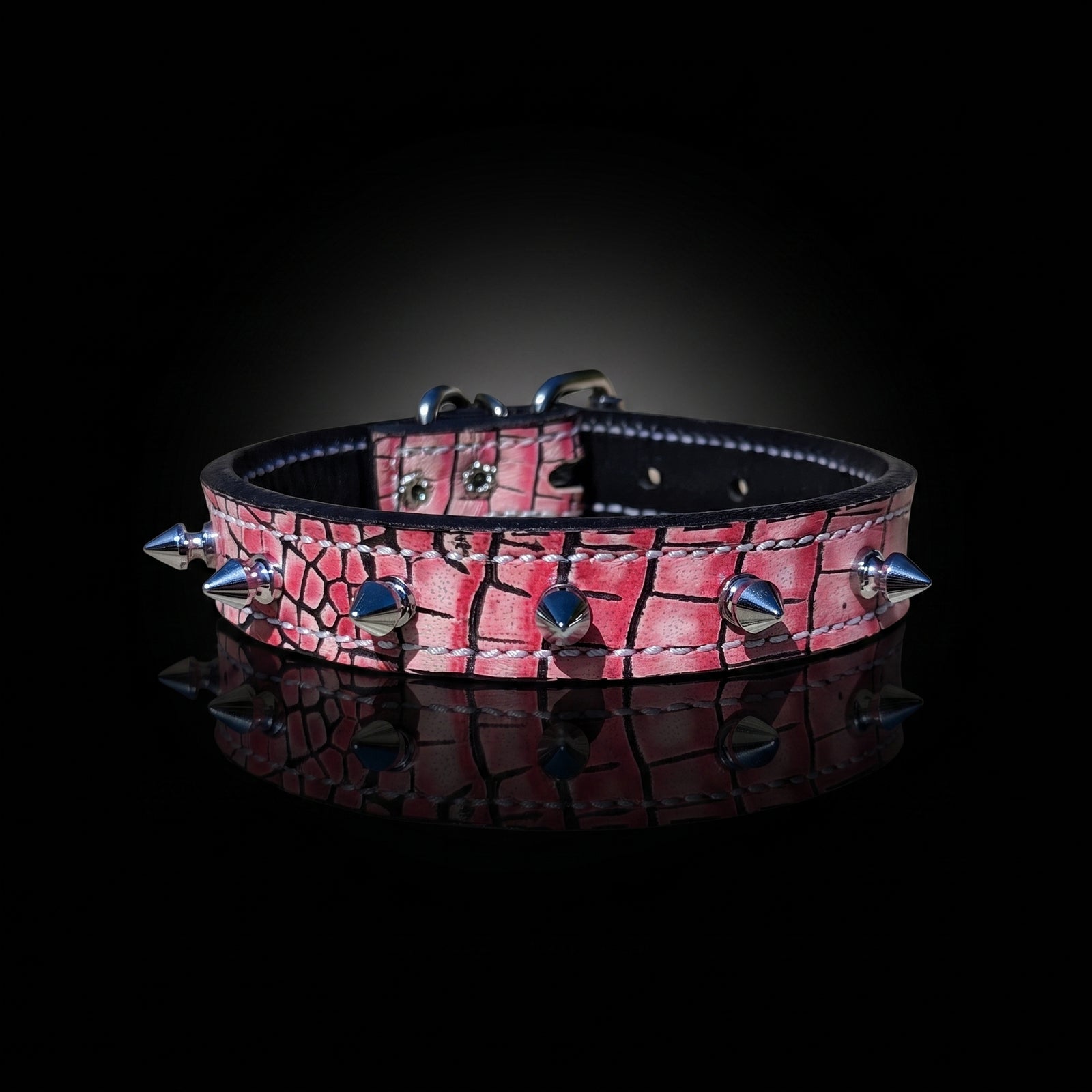 A pink crocodile-embossed leather collar with silver metal spikes and studs, displayed on a reflective black surface against a dark background.