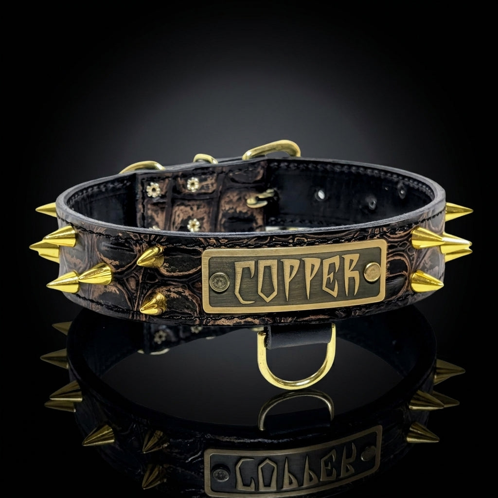 A brown leather spiked collar with gold-toned metal spikes and a central rectangular plate engraved with the word 'COPPER', shown on a reflective black surface against a dark background.