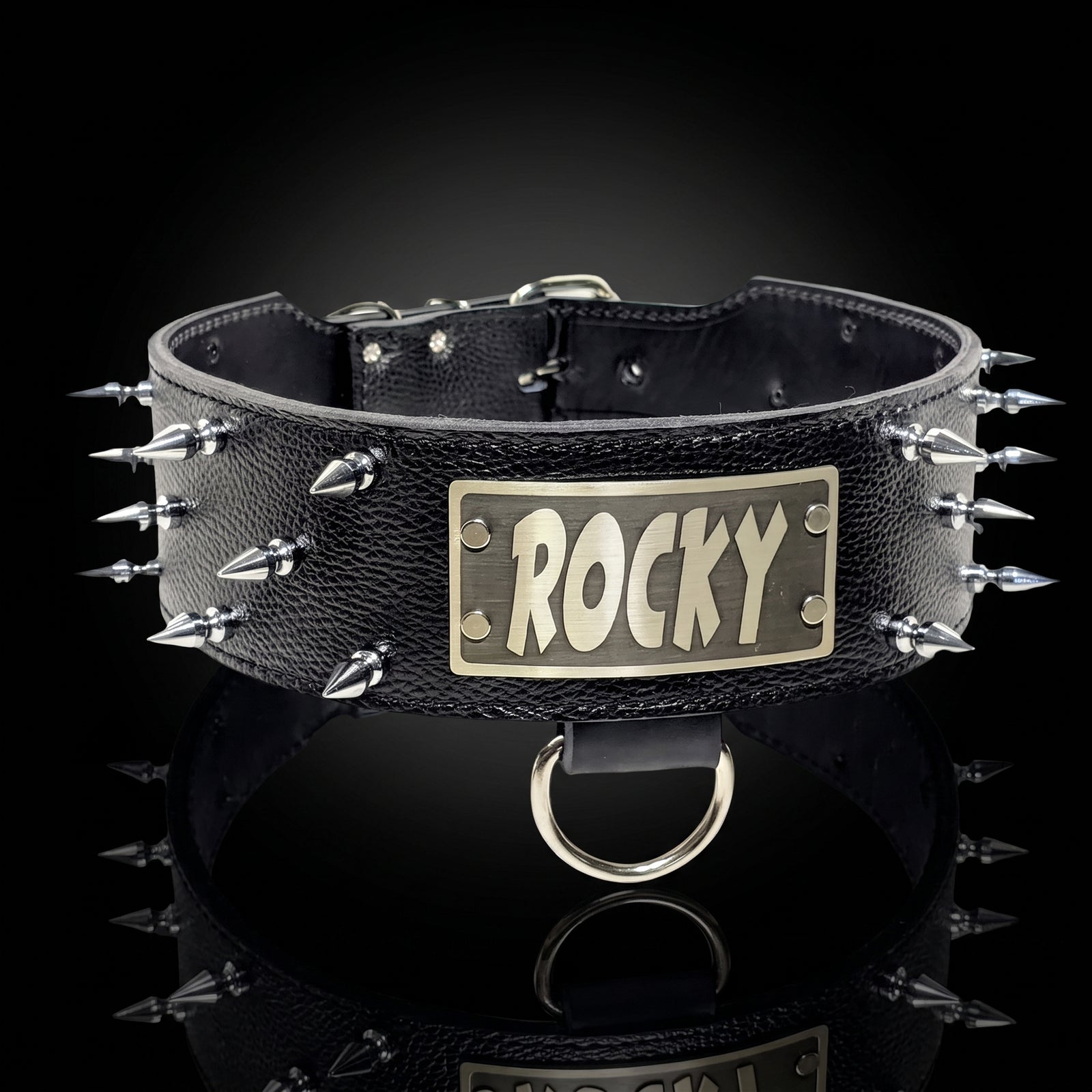 A black textured leather spiked collar with a rectangular brass plate engraved with the name 'ROCKY' in stylized letters, displayed on a reflective black surface against a dark background.