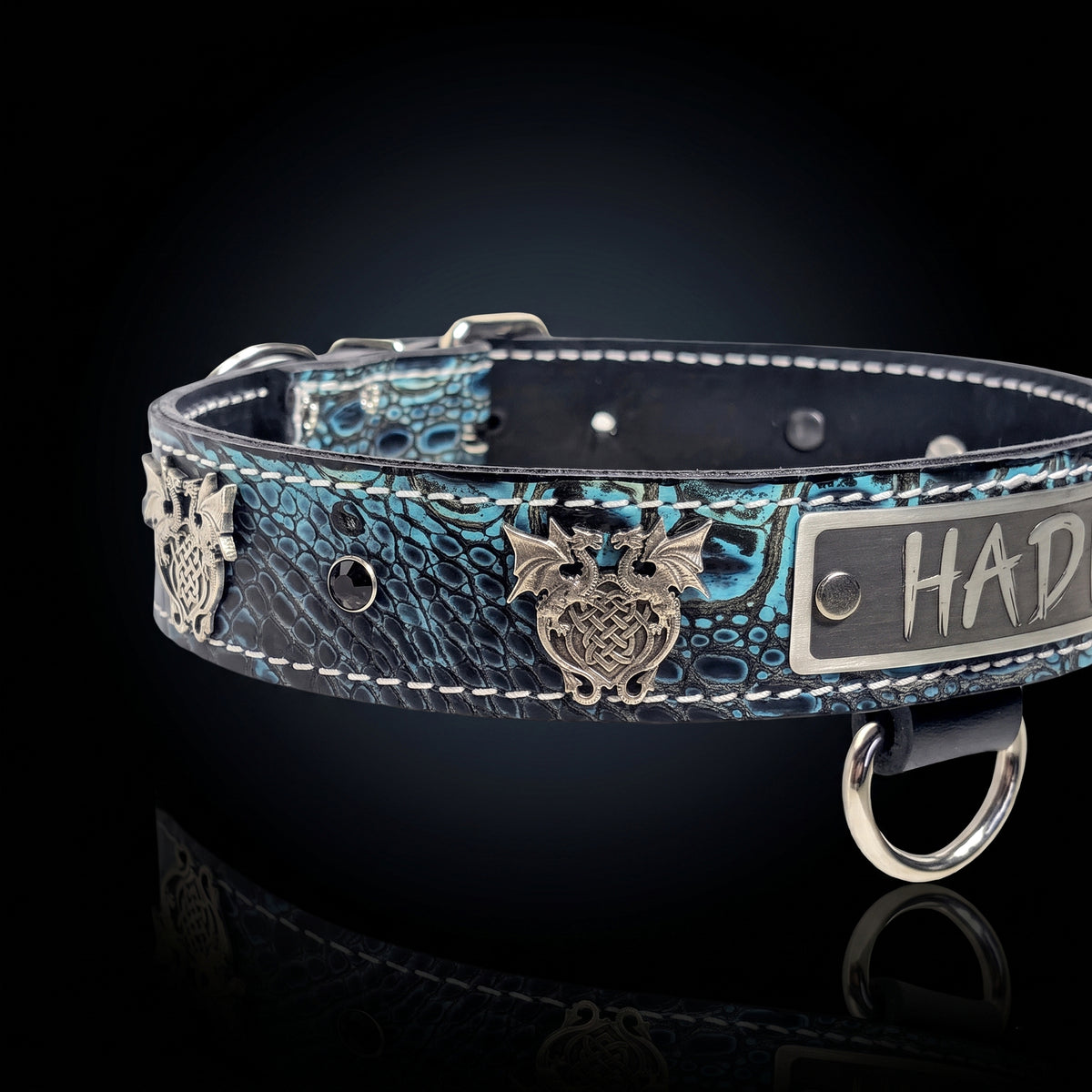 A close-up of a blue and black crocodile-embossed leather collar with silver metal hardware, featuring a central owl emblem and a silver nameplate engraved with 'HAD'.