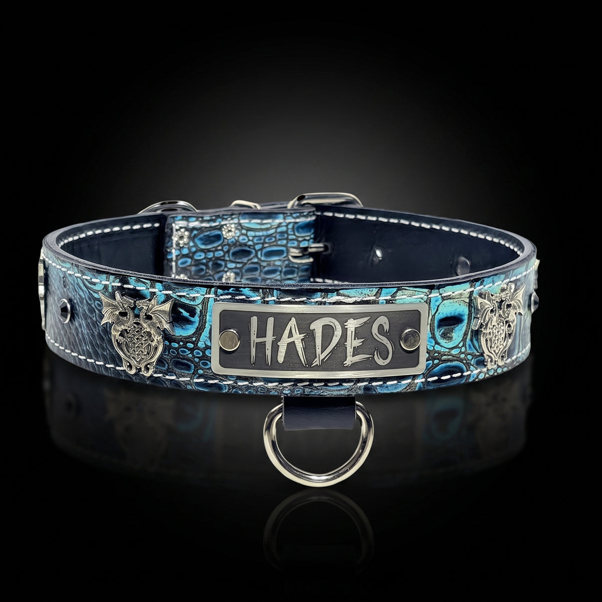 A blue and black crocodile-embossed leather dog collar with silver metal hardware, featuring a central silver plate engraved with the name 'HADES' and decorative owl emblems on both sides, displayed on a reflective black background.