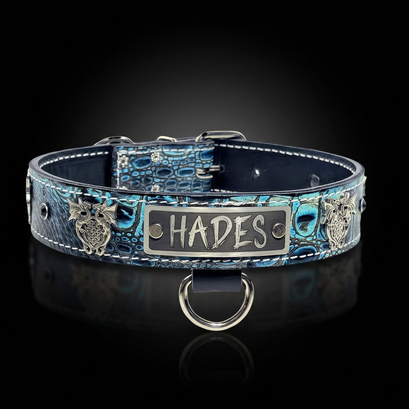 A blue and black crocodile-embossed leather dog collar with silver metal hardware, featuring a central silver plate engraved with the name 'HADES' and decorative owl emblems on both sides, displayed on a reflective black background.