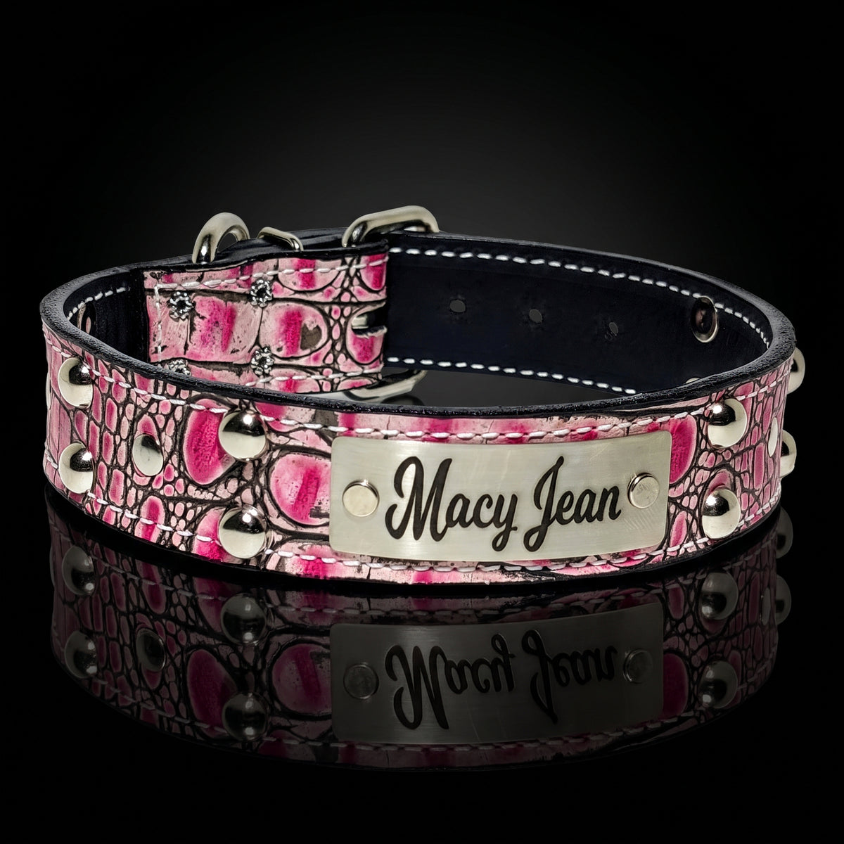 A pink and black crocodile-embossed leather dog collar with silver studs and a silver metal nameplate engraved with 'Macy Jean', shown on a reflective black background.