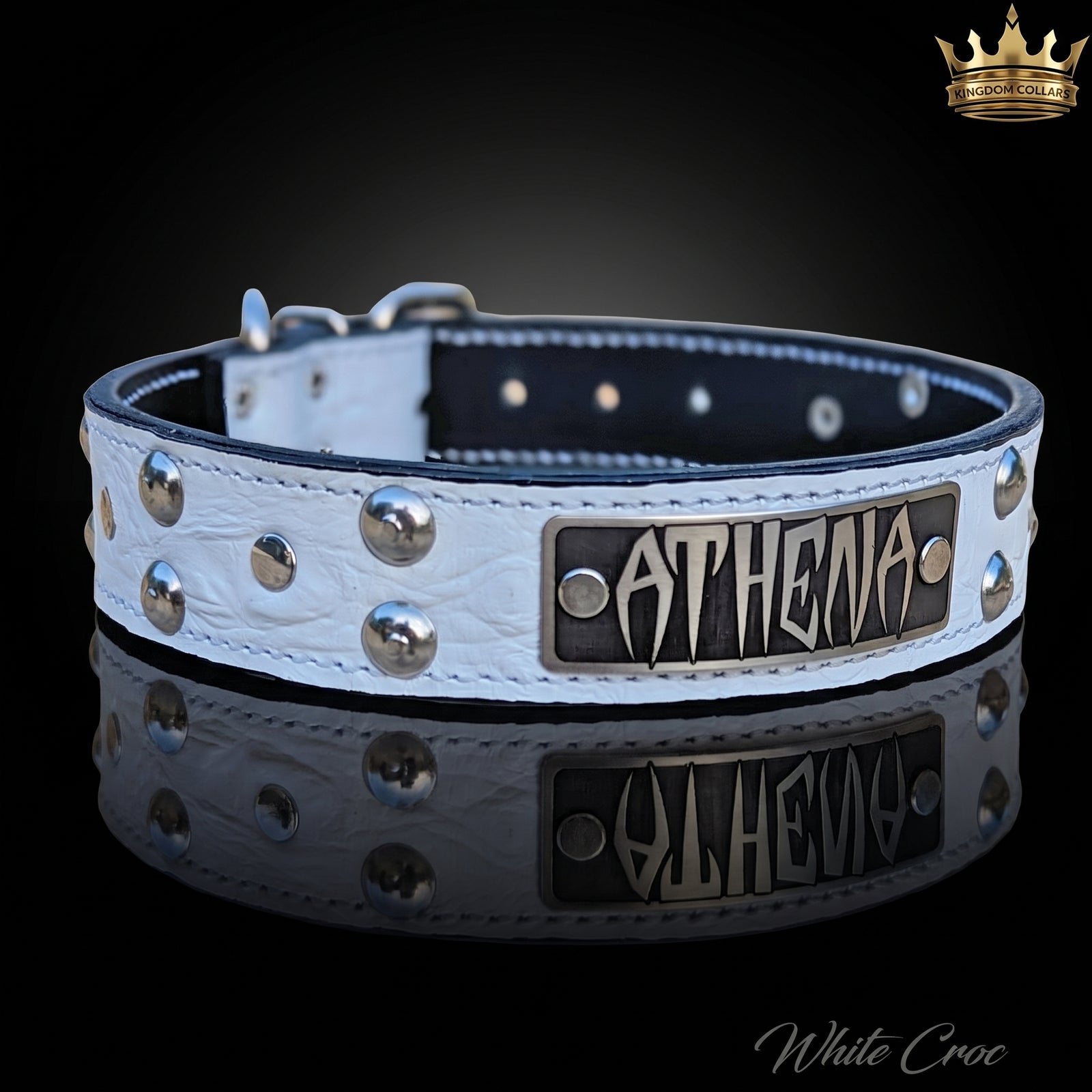 A white crocodile-embossed leather dog collar with silver studs and a black metal plate engraved with the name 'ATHENA', displayed on a reflective black background.
