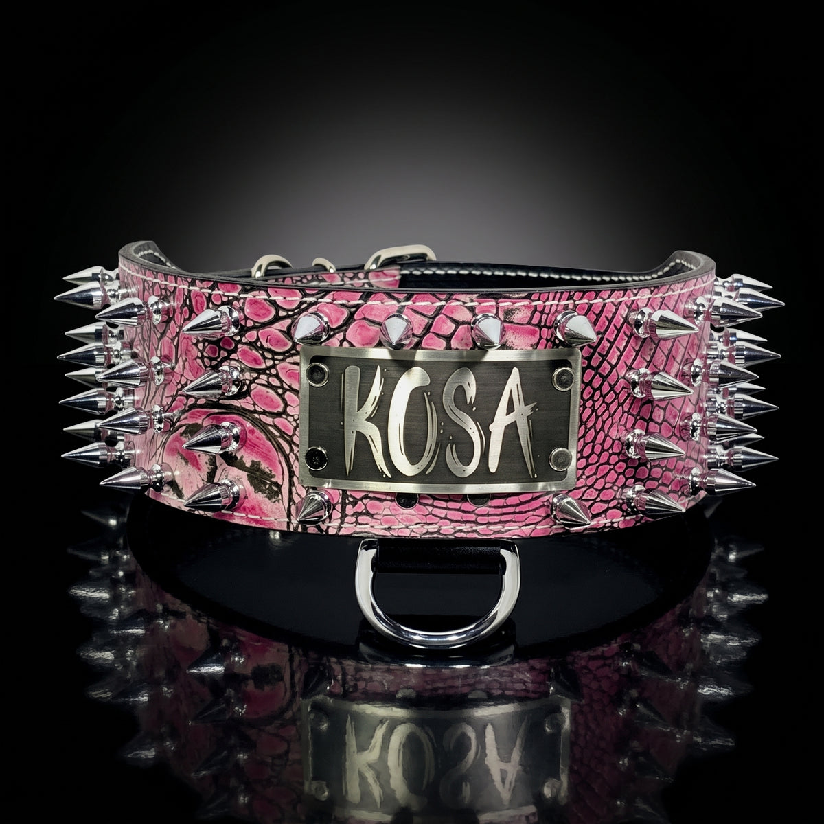 A pink and black croco pattern spiked collar with a silver metal plaque engraved with 'KOSA', displayed on a reflective black surface against a dark background.