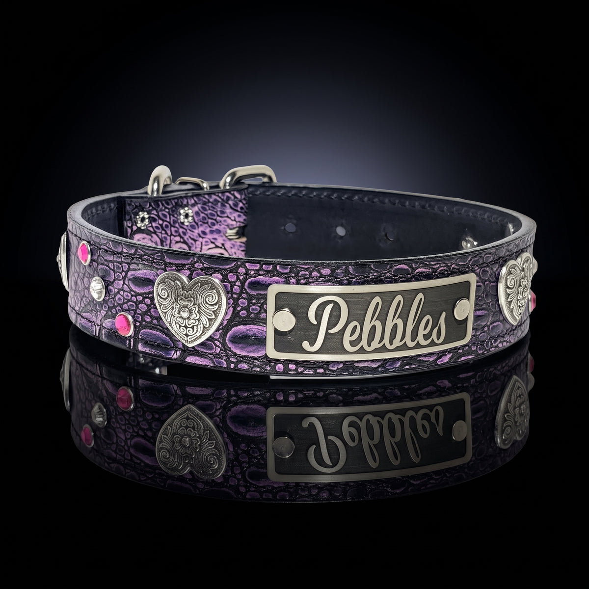 A custom pet collar with a purple and black crocodile-embossed pattern, featuring silver metal accents, a heart-shaped skull emblem, and a rectangular nameplate engraved with the name 'Pebbles'. The collar is displayed on a reflective black surface.